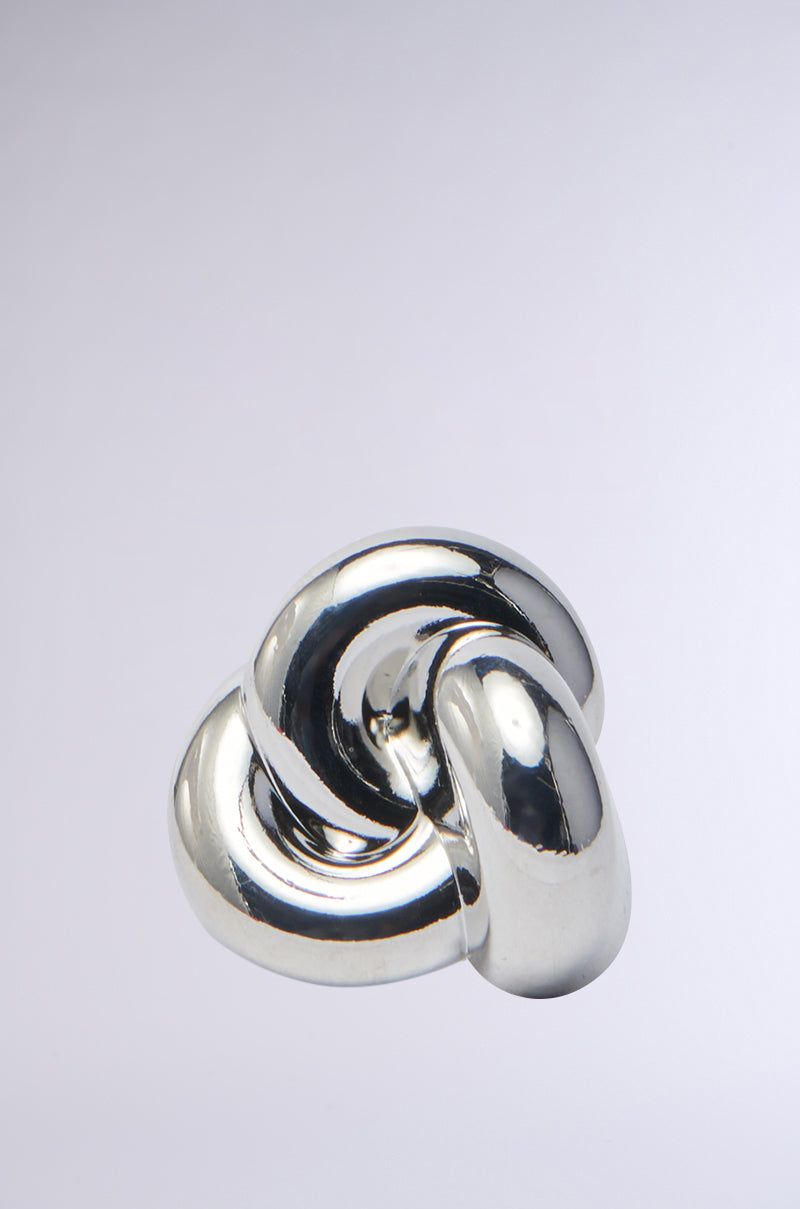 The A LITTLE GOES A LONG WAY SILVER RING features a polished silver knot design with smooth, interlocking loops against a light gray background, reflecting elegantly like a chunky pendant and showcasing its intricate curves.