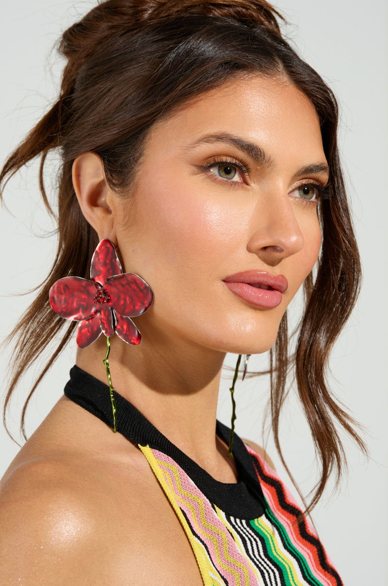 A woman with long brown hair tied back wears large BLOSSOM BEAUTY EARRING featuring floral designs and rhinestone centers, paired with a colorful striped halter top as she gazes to the side against a neutral background. 1