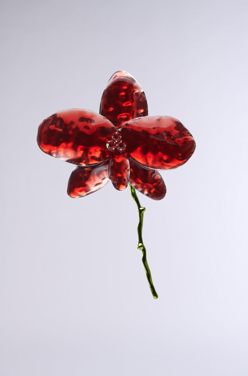 The BLOSSOM BEAUTY EARRING is a glass sculpture of a red orchid with a green stem set on a plain white background. Its glossy petals and rhinestone centers showcase detailed textures and vibrant red hues. 3