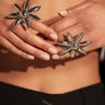 Close-up of hands wearing the IN THE SUN RING SET—two large, metallic flower rings with adjustable bands—posed before a bare midriff. The person sports black pants with a silver ring accent and neatly manicured nails.