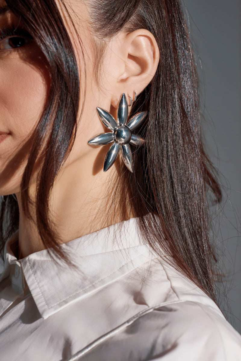Close-up of a woman with long dark hair in a white collared shirt wearing the SARA EARRING—large, flower-shaped faux silver earrings with petals radiating from a central bead.
