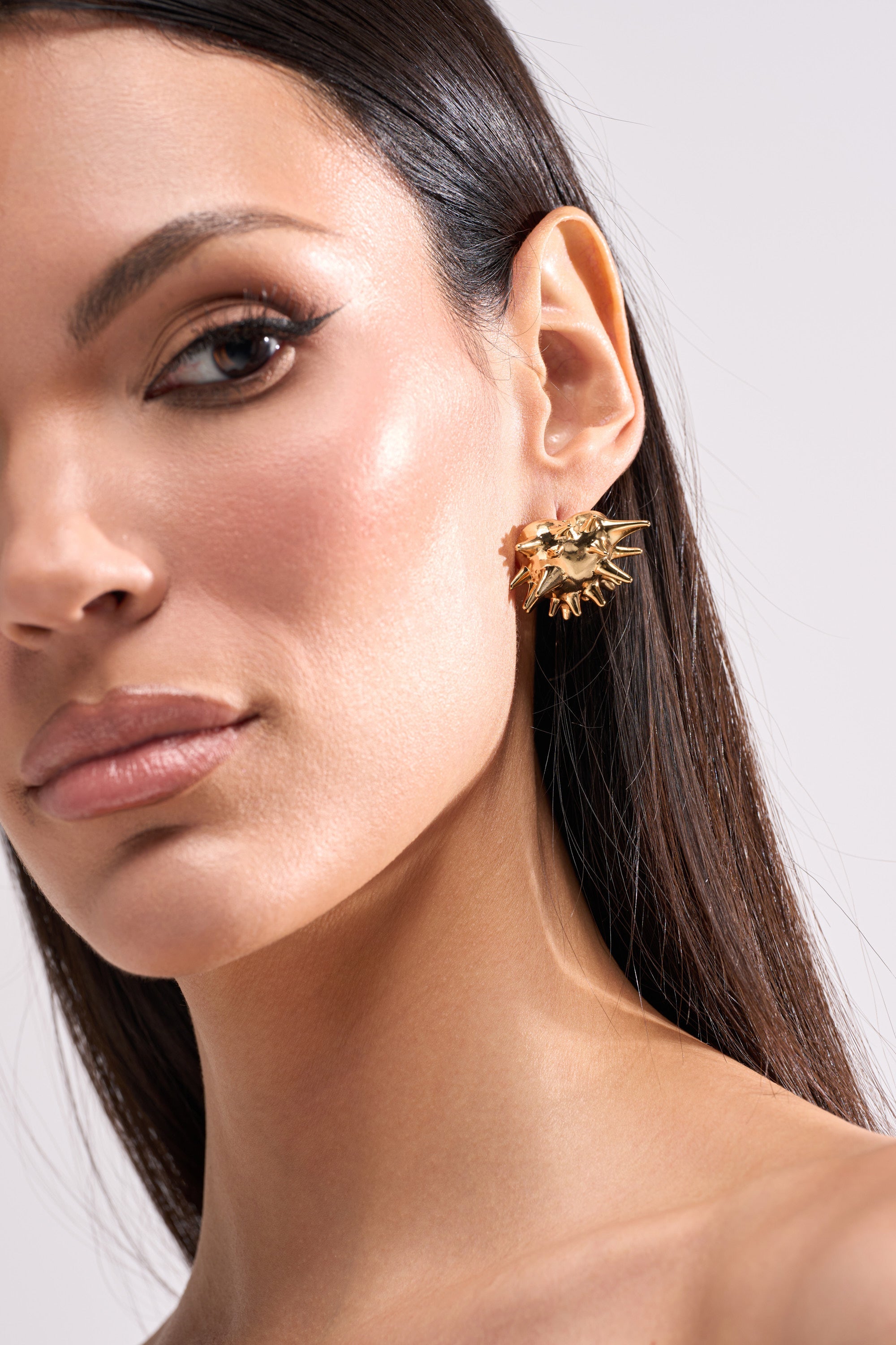 A woman with straight dark hair wears the AKIRA Label TOUCH ME I DARE U EARRING—a bold, gold-tone spiky lion’s head design—shown in close-up from the side against a light background. 1