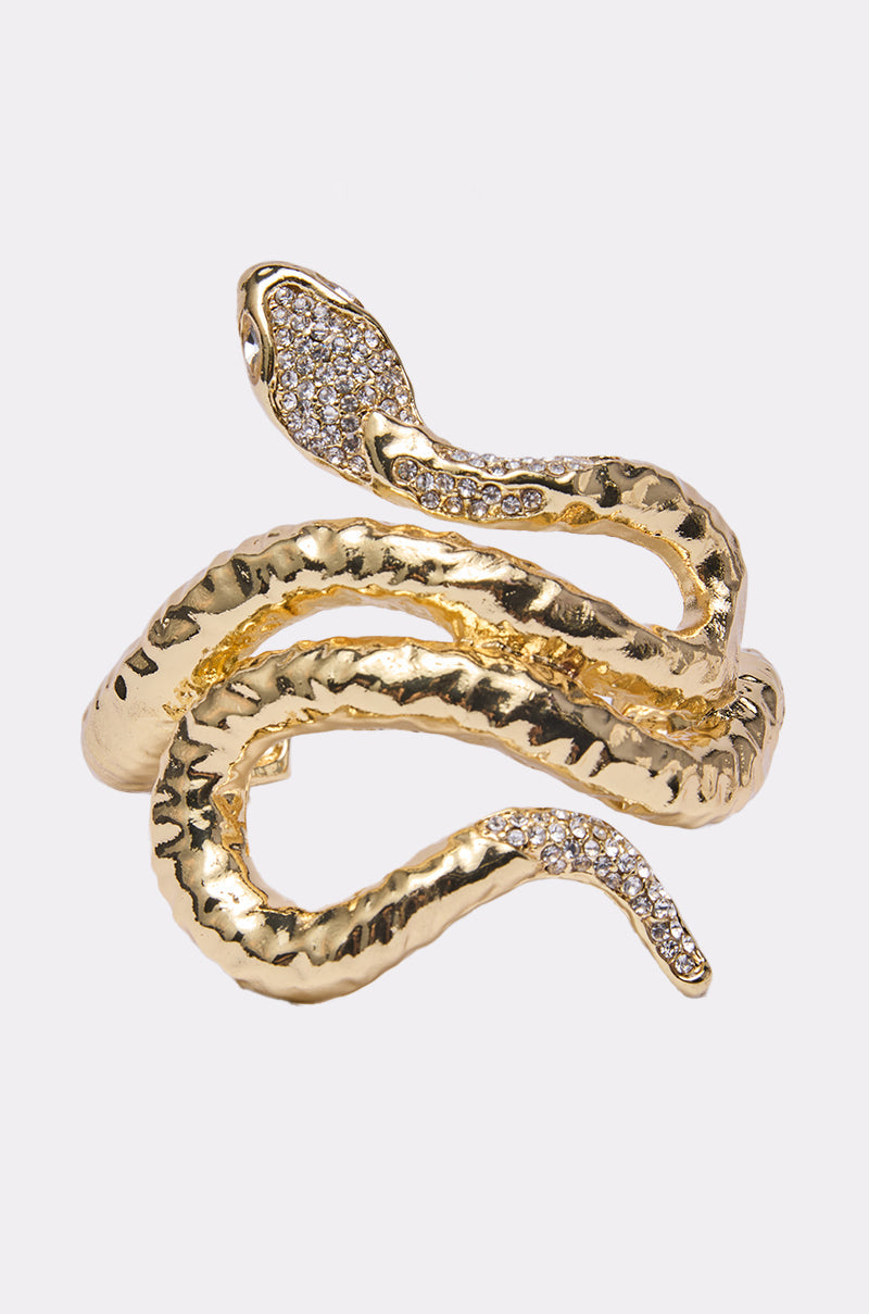 The BITE ME BRACELET is a gold snake cuff shaped like a coiled serpent, with a textured surface and sparkling crystals encrusting the snake’s head and tail.
