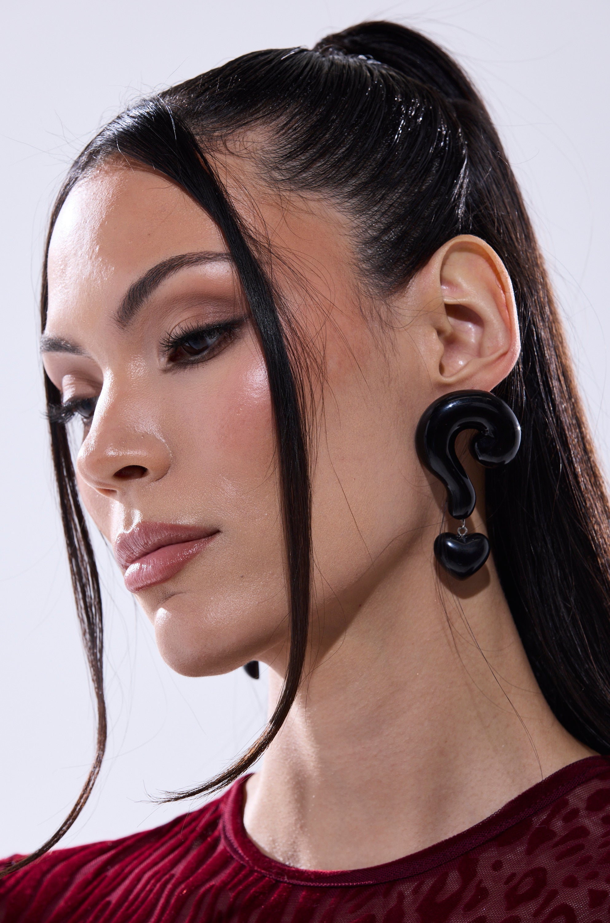 A woman with long, dark hair in a high ponytail wears the NO QUESTIONS EARRING—bold black acrylic statement earrings shaped like question marks—with a burgundy textured top, neutral makeup, and a calm expression as she looks down and away. 3