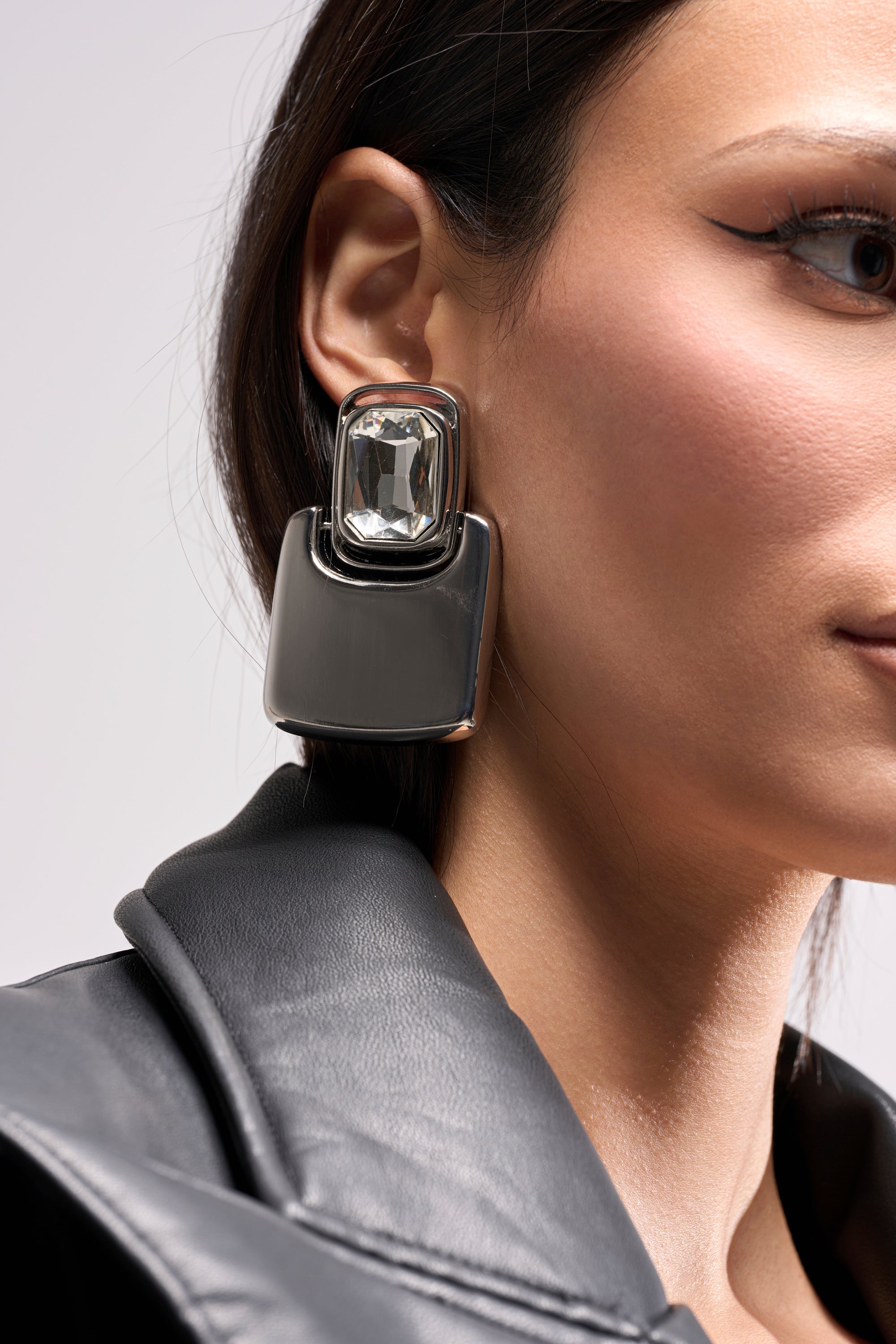 A close-up of a woman in a black leather jacket, winged eyeliner, and striking MOD EARRING—a bold geometric design with a clear gemstone and metallic base. Only part of her face is shown to highlight the earrings. 1
