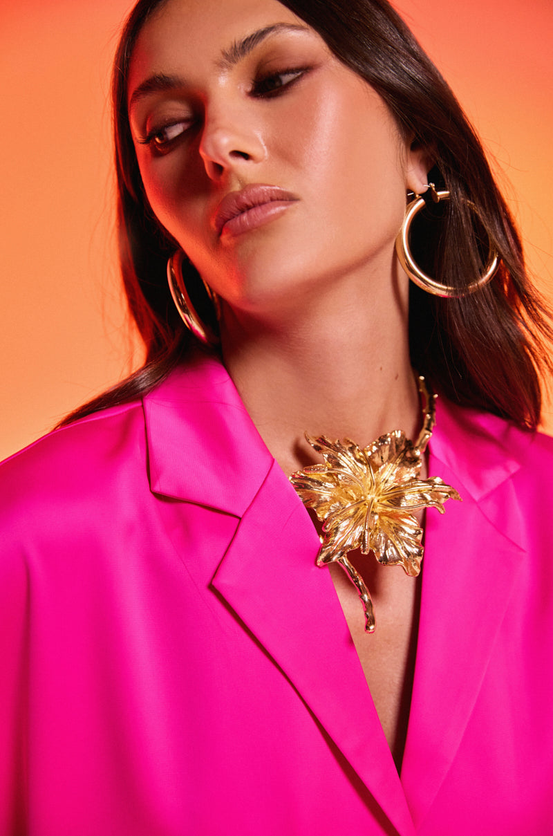 A woman in a hot pink blazer wears large gold hoops and the IM THE ONE FLOWER NECKLACE, featuring an oversized flower motif, posing against an orange gradient background. 1