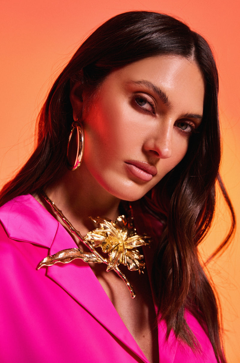 A woman with long brown hair wears the IM THE ONE FLOWER NECKLACE, a bold gold choker with an oversized flower motif, styled with a bright pink blazer and large gold hoop earrings against an orange-pink gradient background. 2