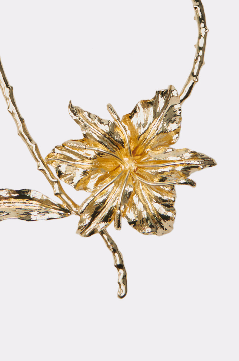 Close-up of the IM THE ONE FLOWER NECKLACE, featuring a gold choker with an oversized flower pendant and textured petals on a stem-like chain, set against a white background. 3