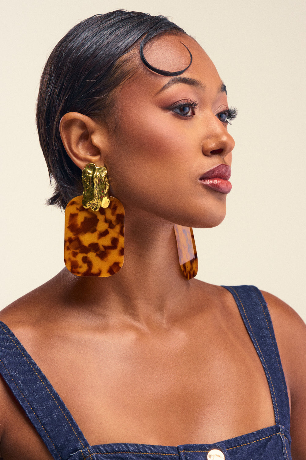 A woman with short, slicked-back hair wears the bold RICH RICH EARRING statement earrings and a chic denim dress, perfectly capturing a retro vibe. Facing right against a plain background, she showcases her striking earrings and flawless makeup.
