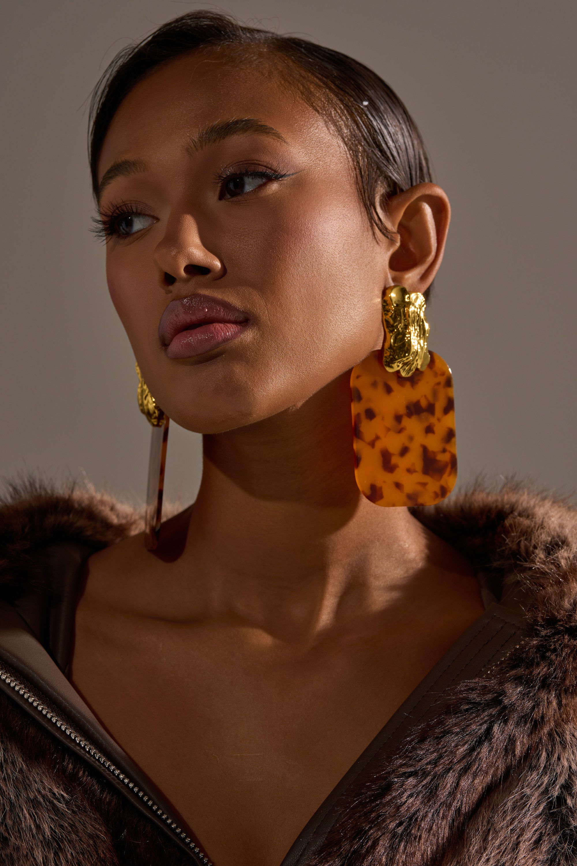 A woman with smooth skin, glossy lips, and dark hair wears RICH RICH EARRING—oversized statement tortoiseshell hoops with gold accents—paired with a brown top and faux fur coat. She gazes upward against a plain backdrop.