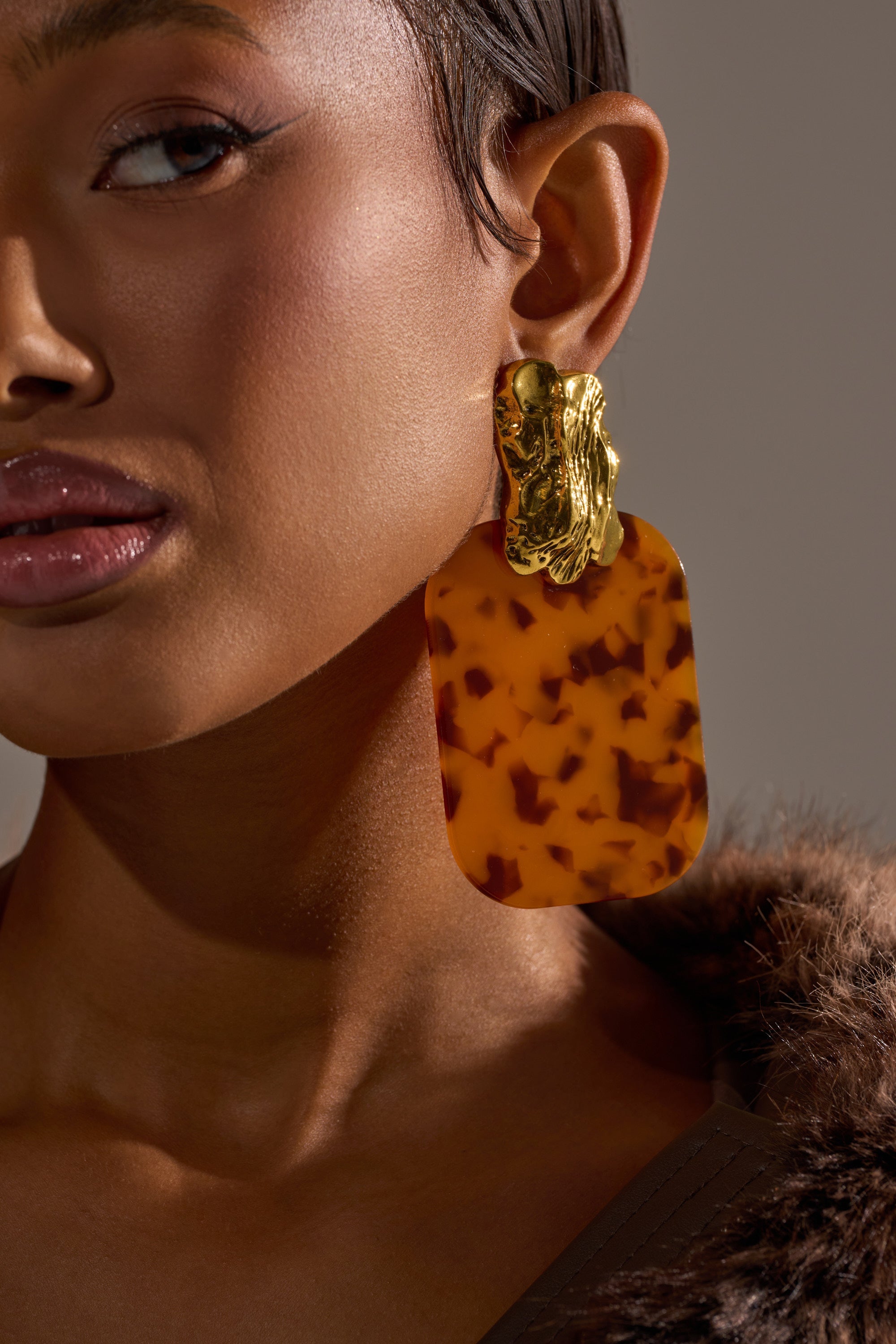 Close-up of a woman wearing the RICH RICH EARRING, featuring bold gold-tone studs and rectangular tortoiseshell acrylic dangles. She has smooth skin, glossy lips, and is dressed in a brown faux fur garment. 1
