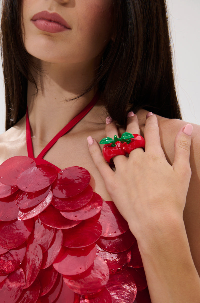 A woman with long brown hair wears a halter top covered in large, shiny red sequins and the CHERRY BOMB RING, a bold statement accessory with green cherry-shaped accents. Her hand rests on her chest, nails painted light pink.