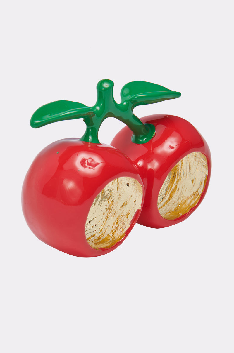 The CHERRY BOMB RING features two shiny red cherries with green stems and leaves, each cherry accented by a gold hollowed-out side—making it a bold accessory for any look or a striking double-finger ring.