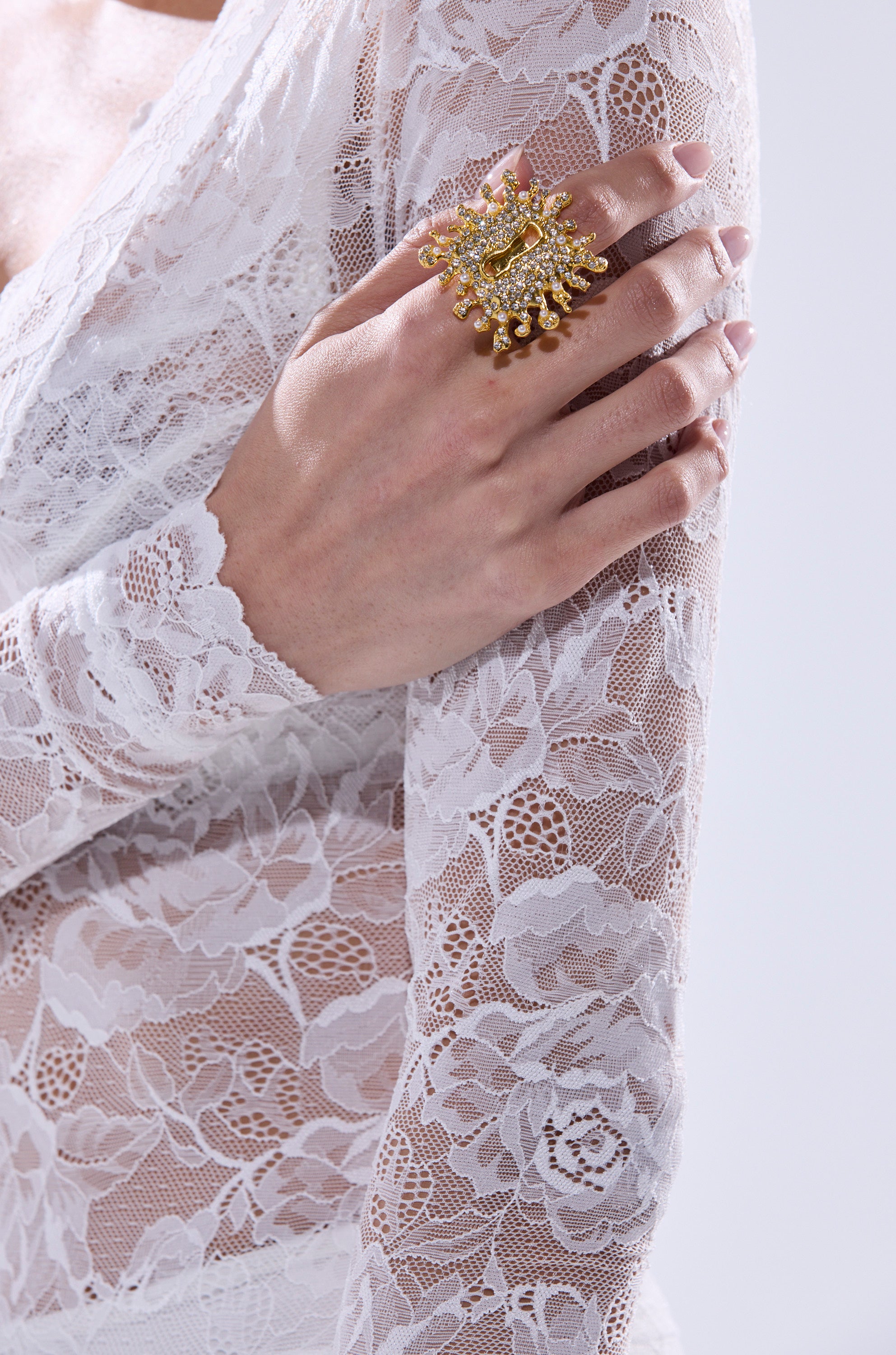 Wearing a white lace long-sleeve top, someone rests their hand on their upper arm, highlighting the UNLOCK ME RING—a striking gold accessory featuring intricate details and dangling accents. 1