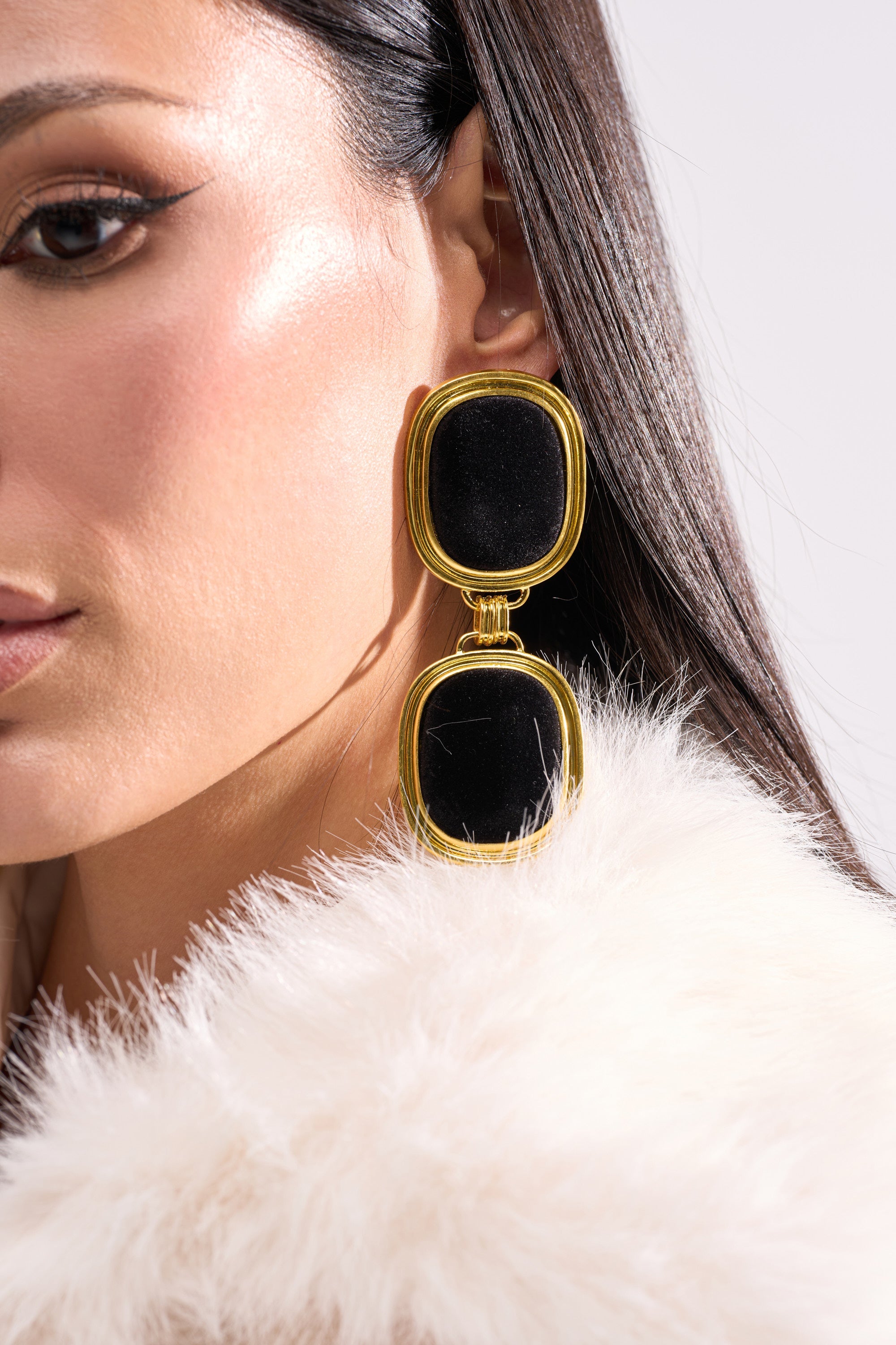 Close-up of a woman with straight dark hair, winged eyeliner, glowing skin, white faux fur, and dramatic black and gold ART DECO EARRING. 1