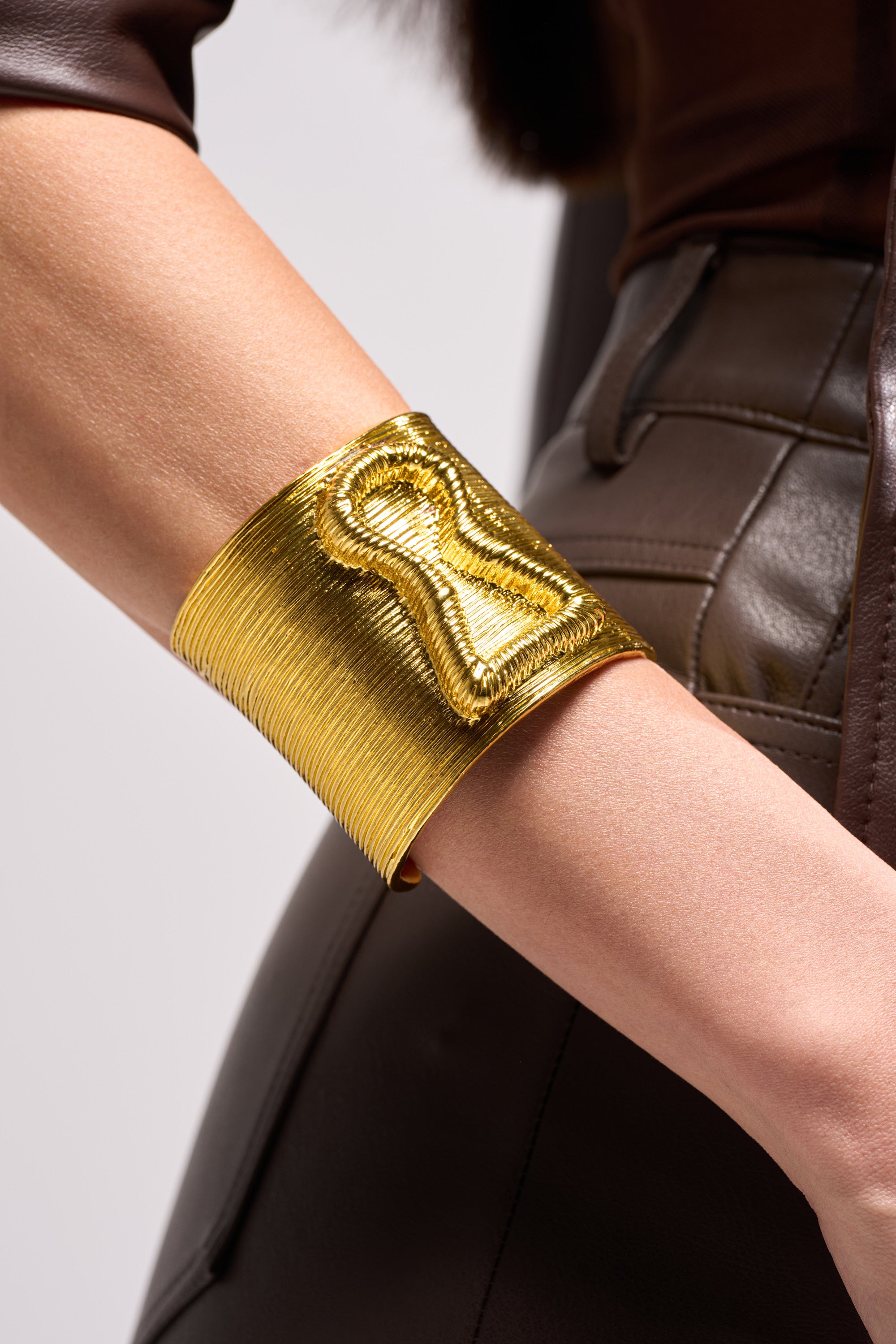 A close-up of a person wearing the IM THE KEY BRACELET, a wide textured metallic piece on their forearm, paired with a brown leather-like outfit featuring visible stitching.