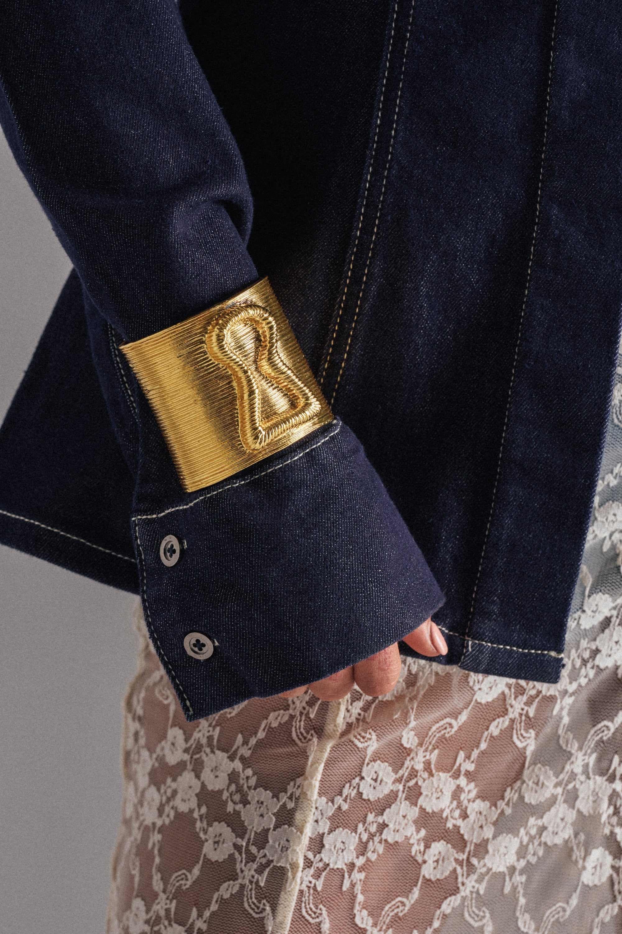 A close-up of a person's arm wearing a dark denim jacket with keyhole embroidery on the cuff, styled with the IM THE KEY BRACELET and a sheer white lace skirt. 0
