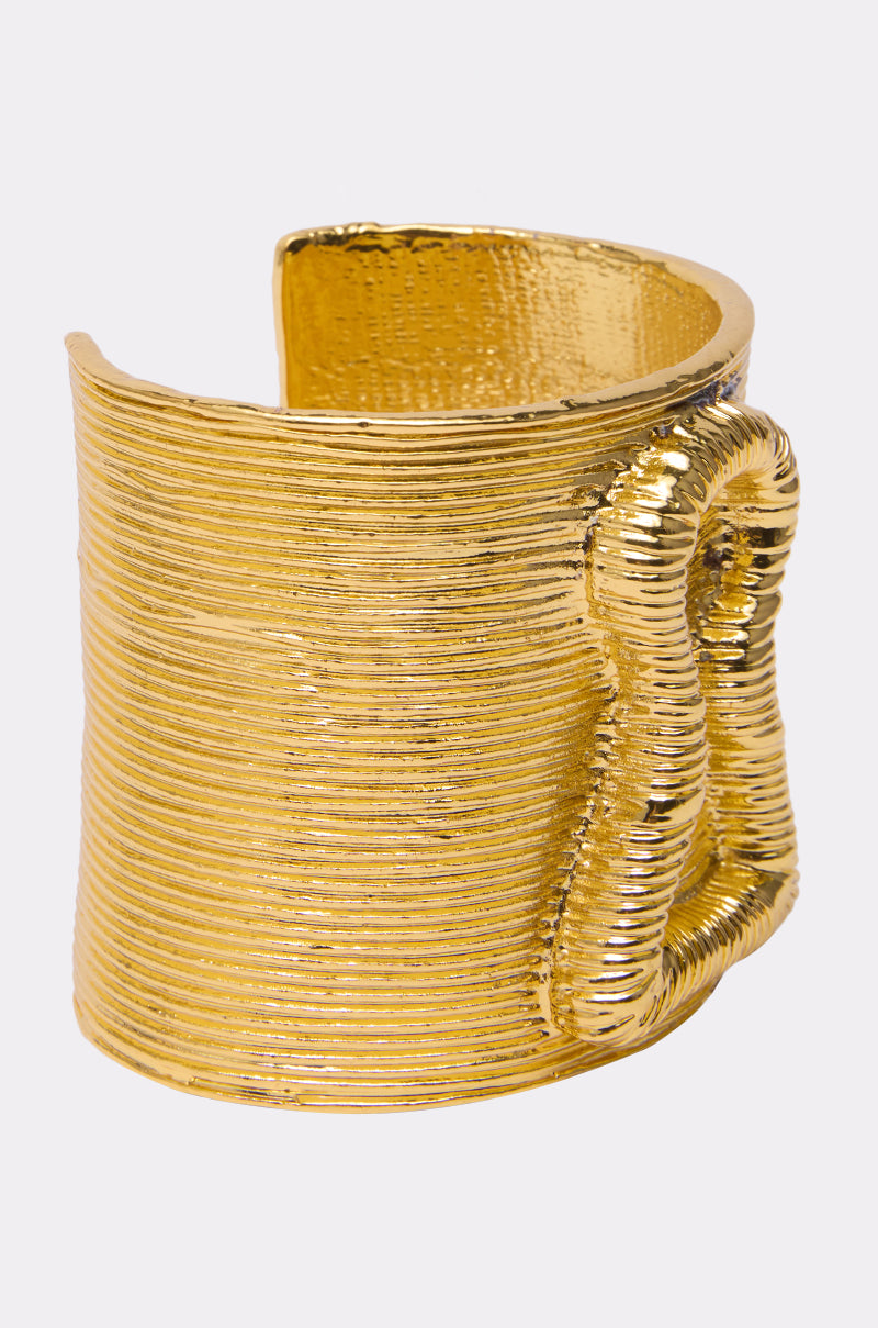 The IM THE KEY BRACELET is a wide, textured gold cuff featuring a wavy, looped design on one side, making it a striking statement piece that stands out against a plain light background.