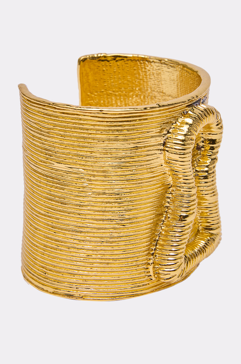 The IM THE KEY BRACELET features a bold gold cuff with horizontal ridges and an abstract looping design on one side, making it a striking statement piece against a light gray background.