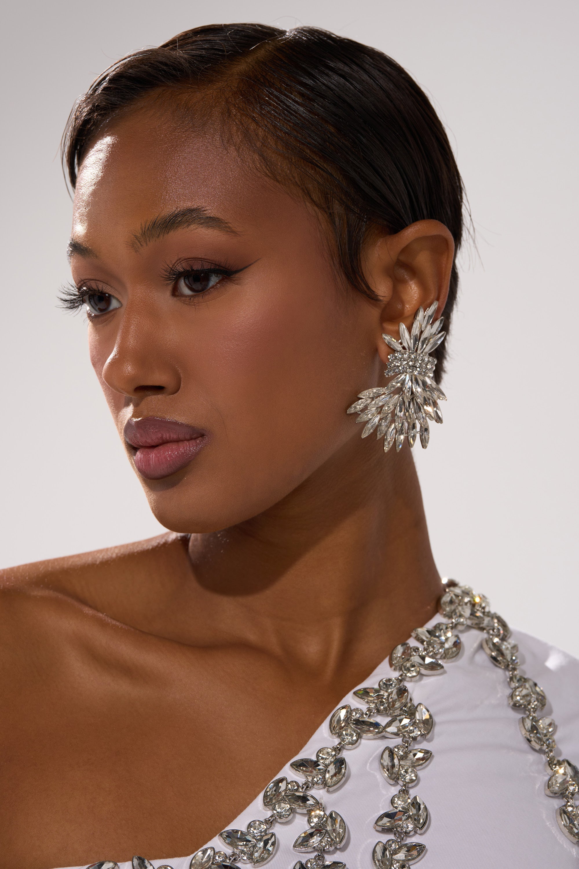 A woman with smooth brown skin and short, sleek hair wears the sparkling, starburst-shaped CICI EARRING and a white, one-shoulder embellished dress, looking slightly down and to the left against a plain background. 1