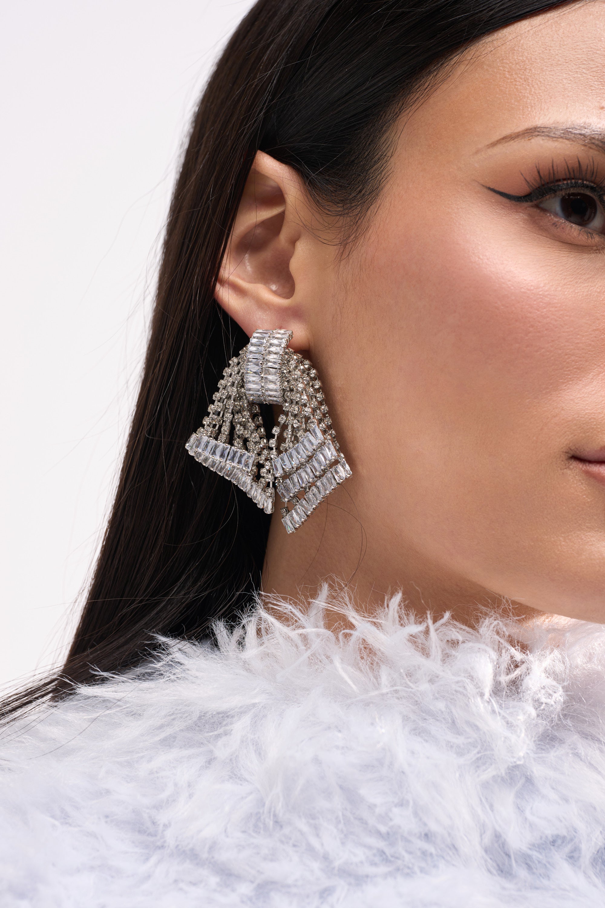 A close-up of a woman wearing the bold ELI EARRING, featuring cascading crystal geometric tiers, with straight dark hair, winged eyeliner, and a white feathery top.