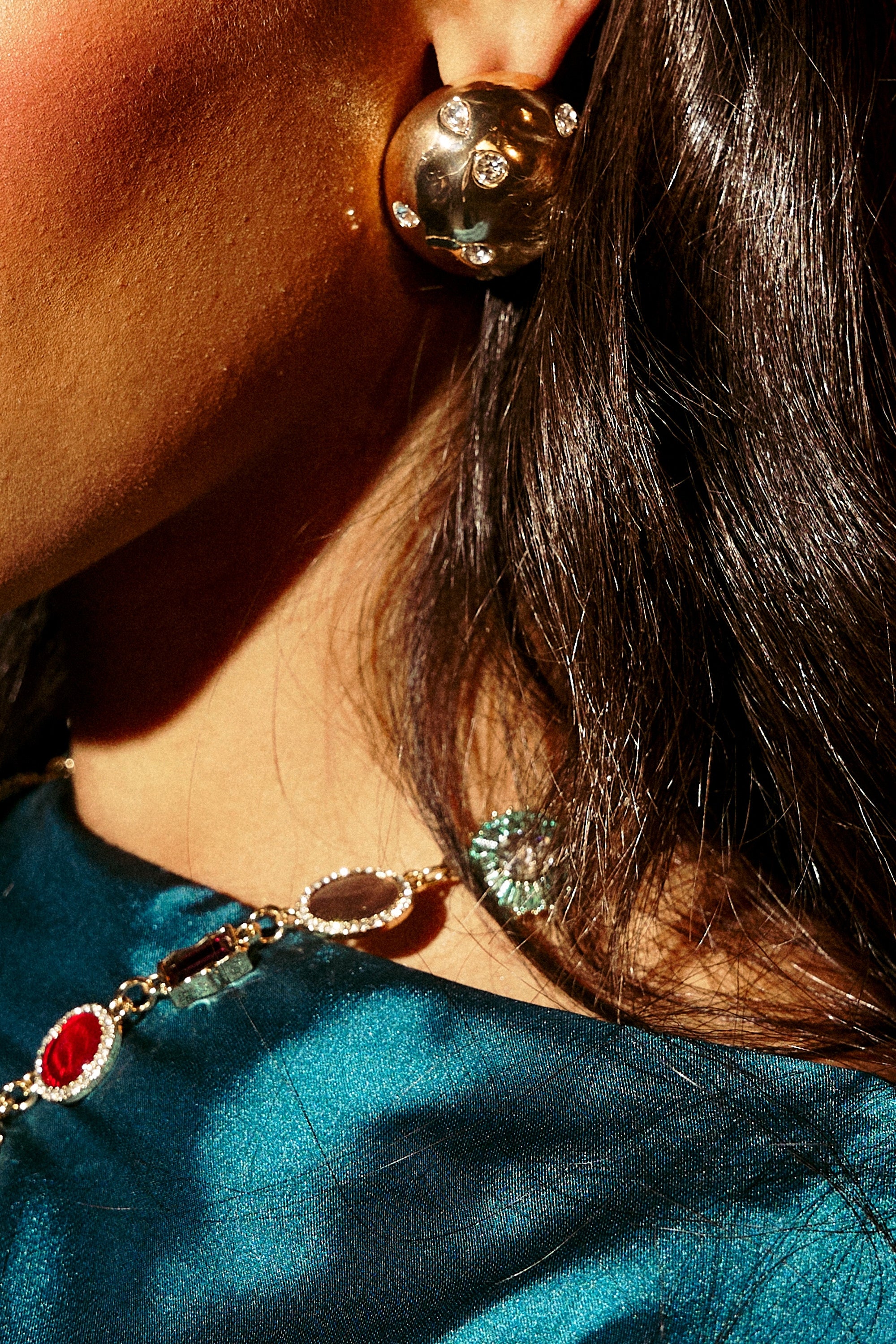 Close-up of a woman's ear wearing PARI EARRING, featuring glittering gold rhinestones with embedded stones, paired with a multicolored gemstone necklace and a shiny blue-green top. Her dark hair is tucked behind her ear. 0
