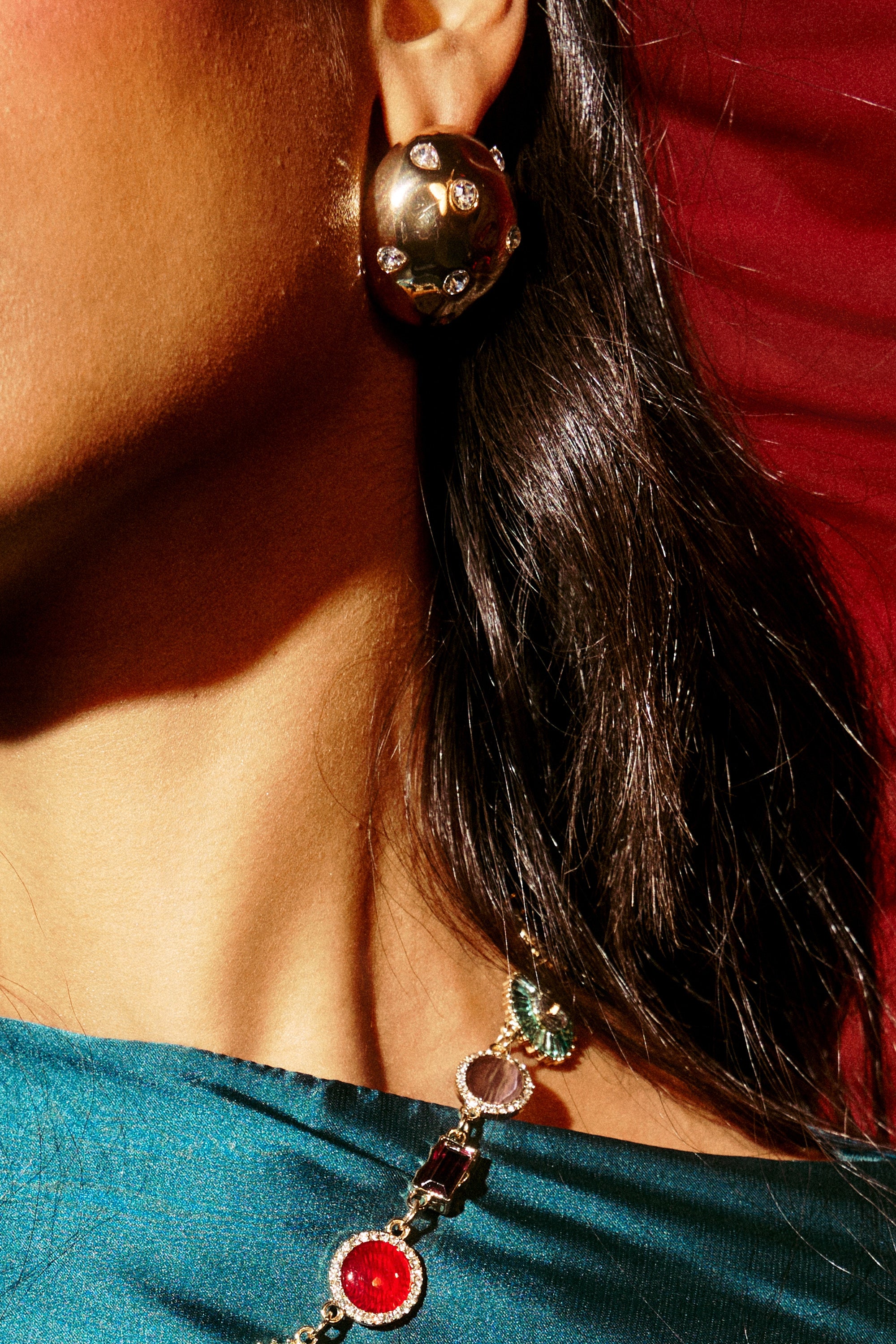 Close-up of a person with long, dark hair in teal fabric wearing PARI EARRING gold metallic earrings adorned with rhinestones and a colorful beaded necklace, set against a red background. 1