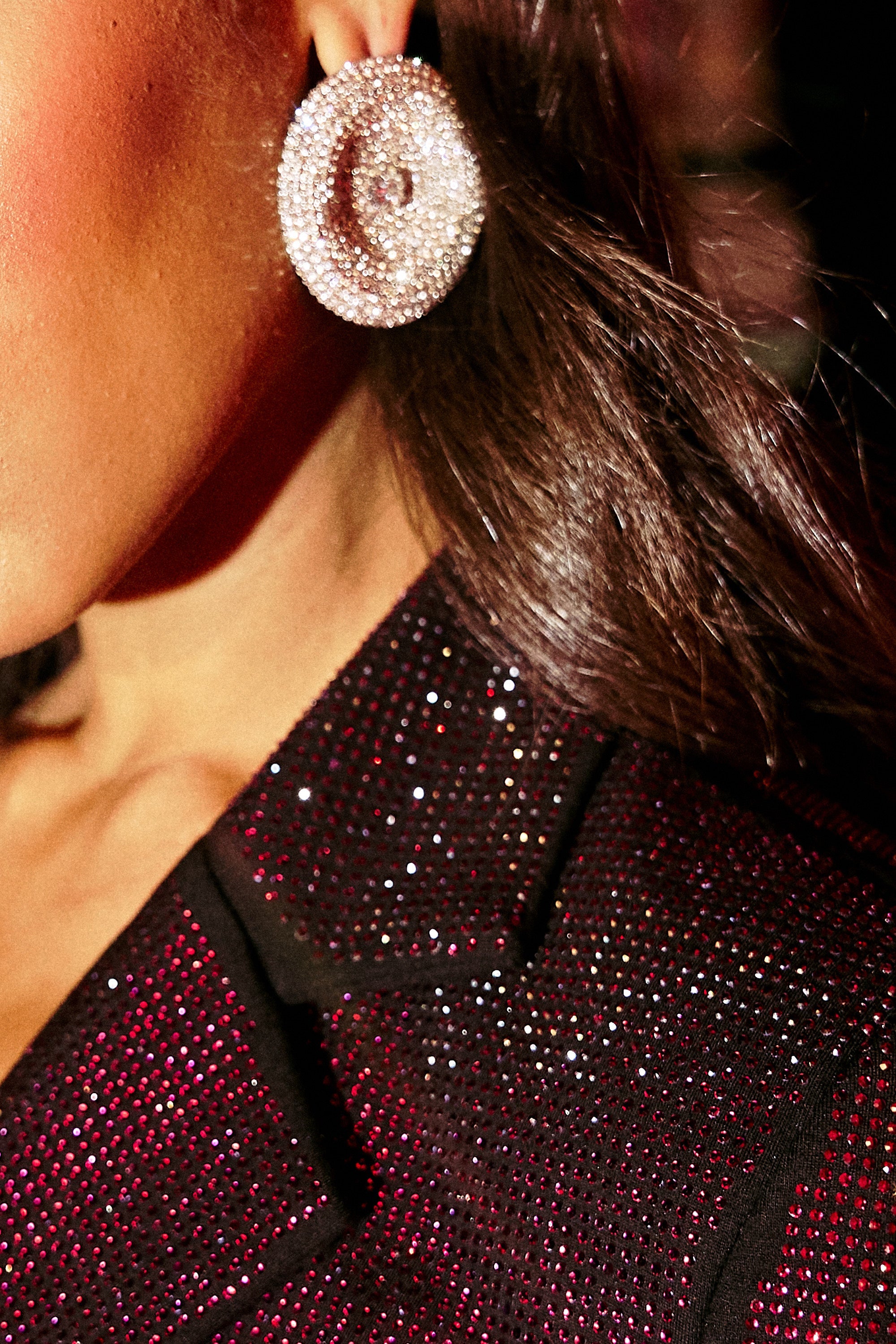 Close-up of a person wearing oversized U WISH EARRING and a sparkly, dark blazer. Only part of the face, ear, and hair are visible, highlighting the blazer's glittery texture and the dazzling accessory. 1