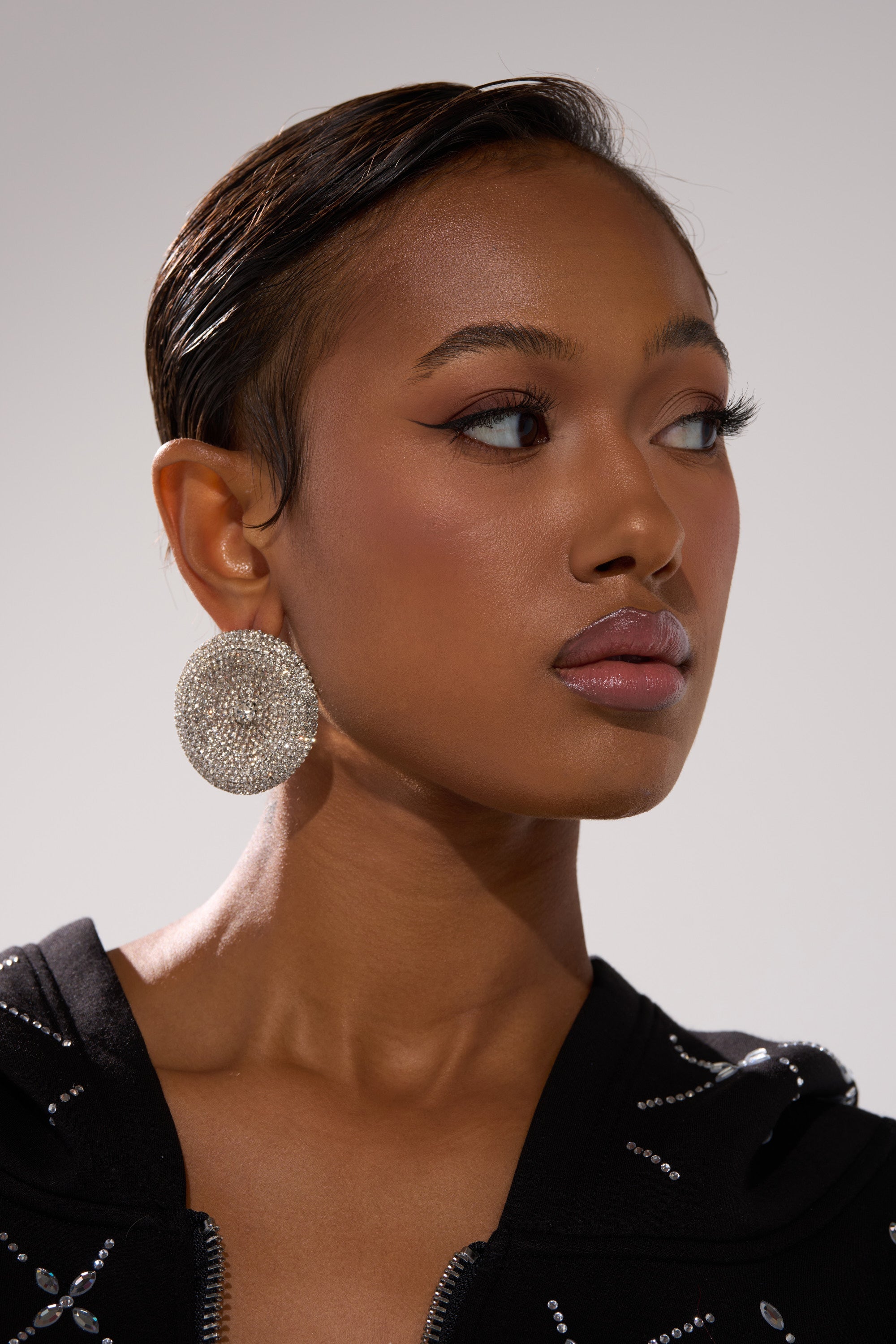 A woman with smooth, dark skin and sleek, pulled-back hair wears the bold U WISH EARRING and a black top with silver details, looking to the side with a calm, confident expression. 3