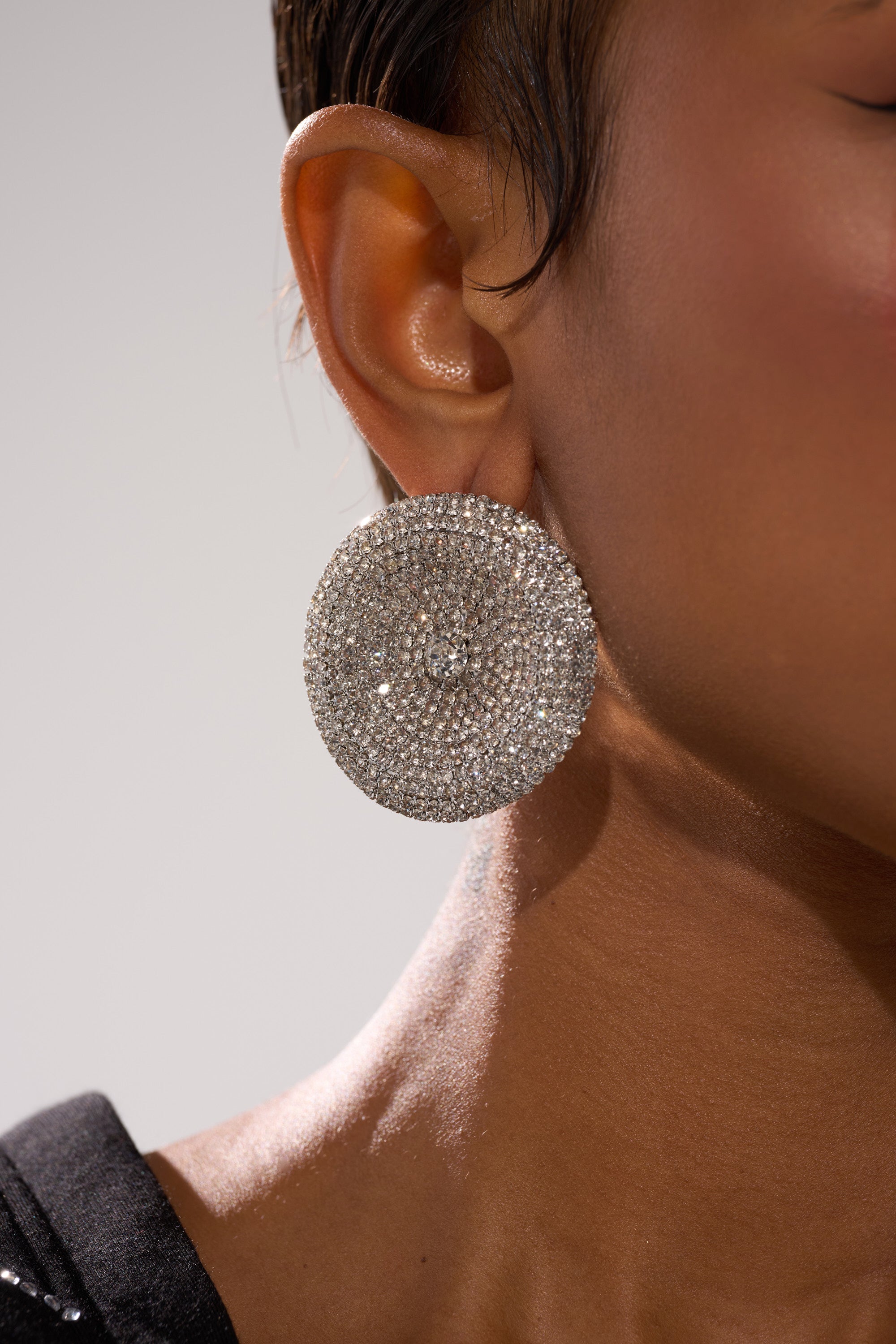 Close-up of a person’s ear wearing the U WISH EARRING, a large, round, silver rhinestone piece. The individual has short hair and smooth skin; the image highlights the earring and ear against a neutral background.