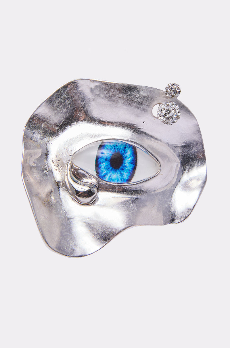 The EYE SEE YOU RING is a silver abstract ring shaped like an eye, featuring a realistic blue iris at the center, rhinestone accents, and two small round gemstones above it. 0