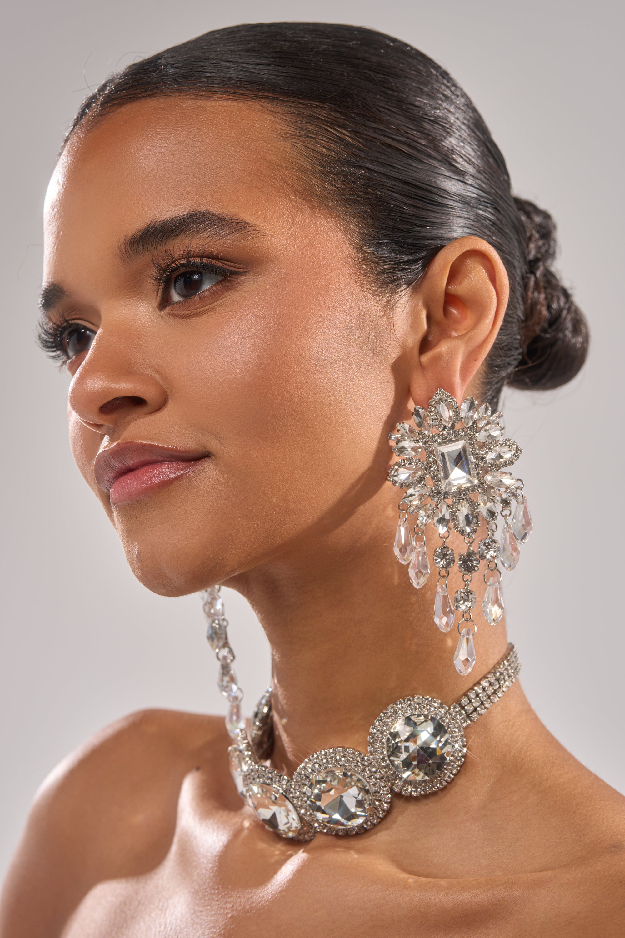 A woman with smooth, dark hair in a low bun wears THE MAIN MOMENT EARRING, adding sparkle to her polished look. Her makeup has a subtle glow as she gazes slightly to the side against a neutral background.