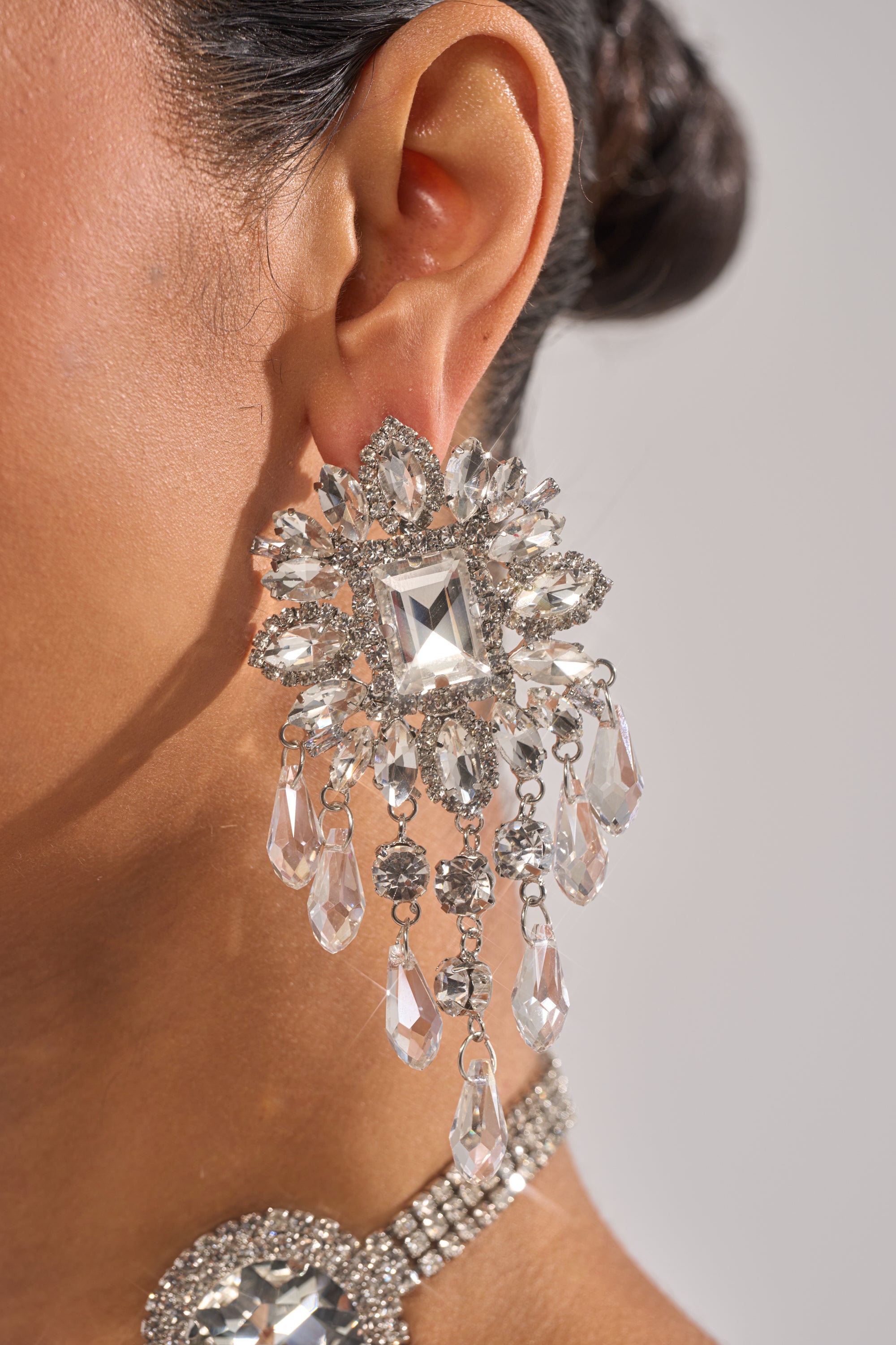 A close-up of a woman's ear wearing THE MAIN MOMENT EARRING, featuring large chandelier-style rhinestones in cascading teardrop shapes. Her hair is styled in an updo, showcasing the sparkling statement piece.