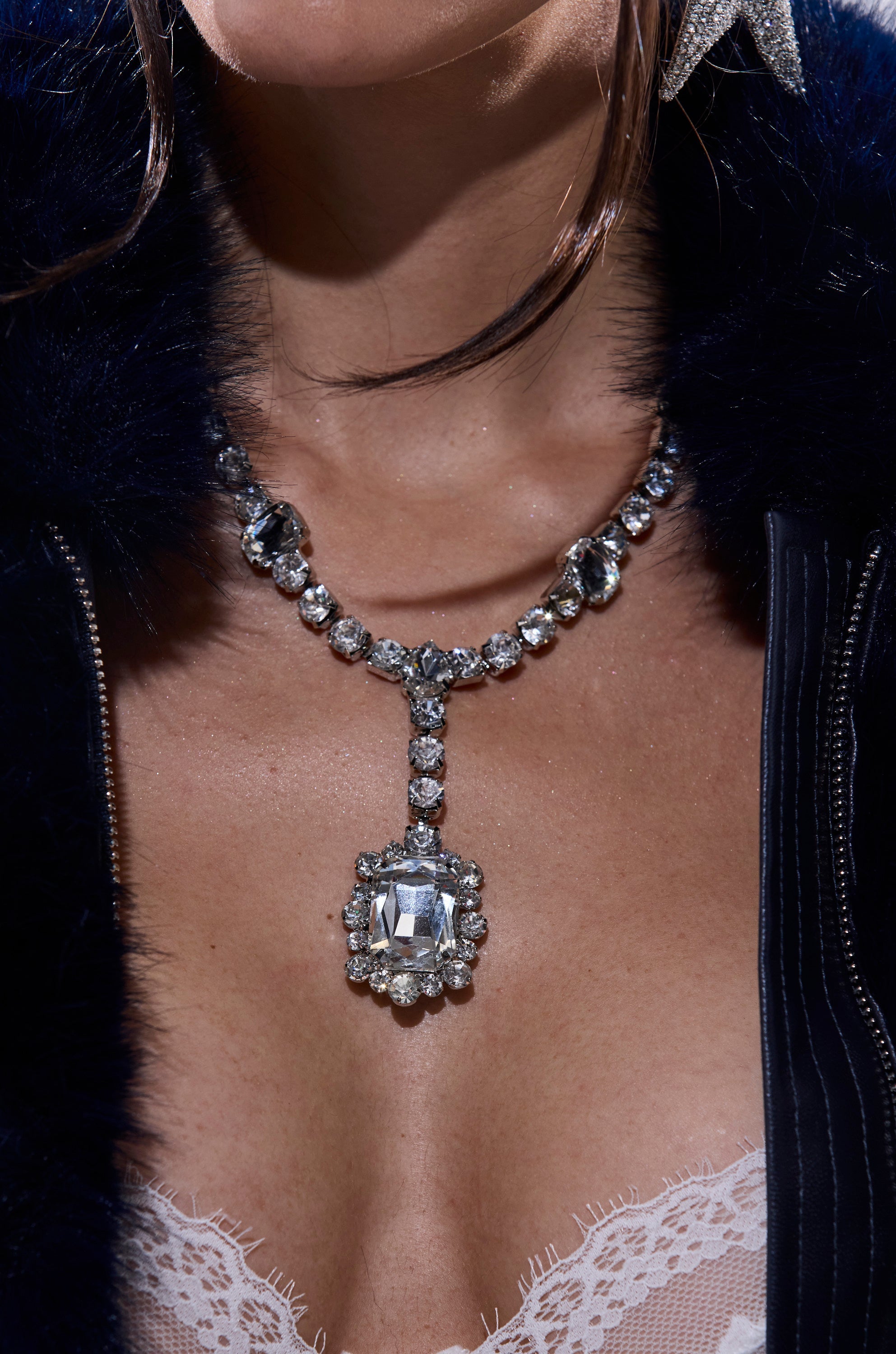 A close-up of a woman in a navy fur coat and white lace bra, her brown hair framing her face, wearing the FOR THE RECORD BLING NECKLACE with a sparkling central crystal rhinestone pendant. 1