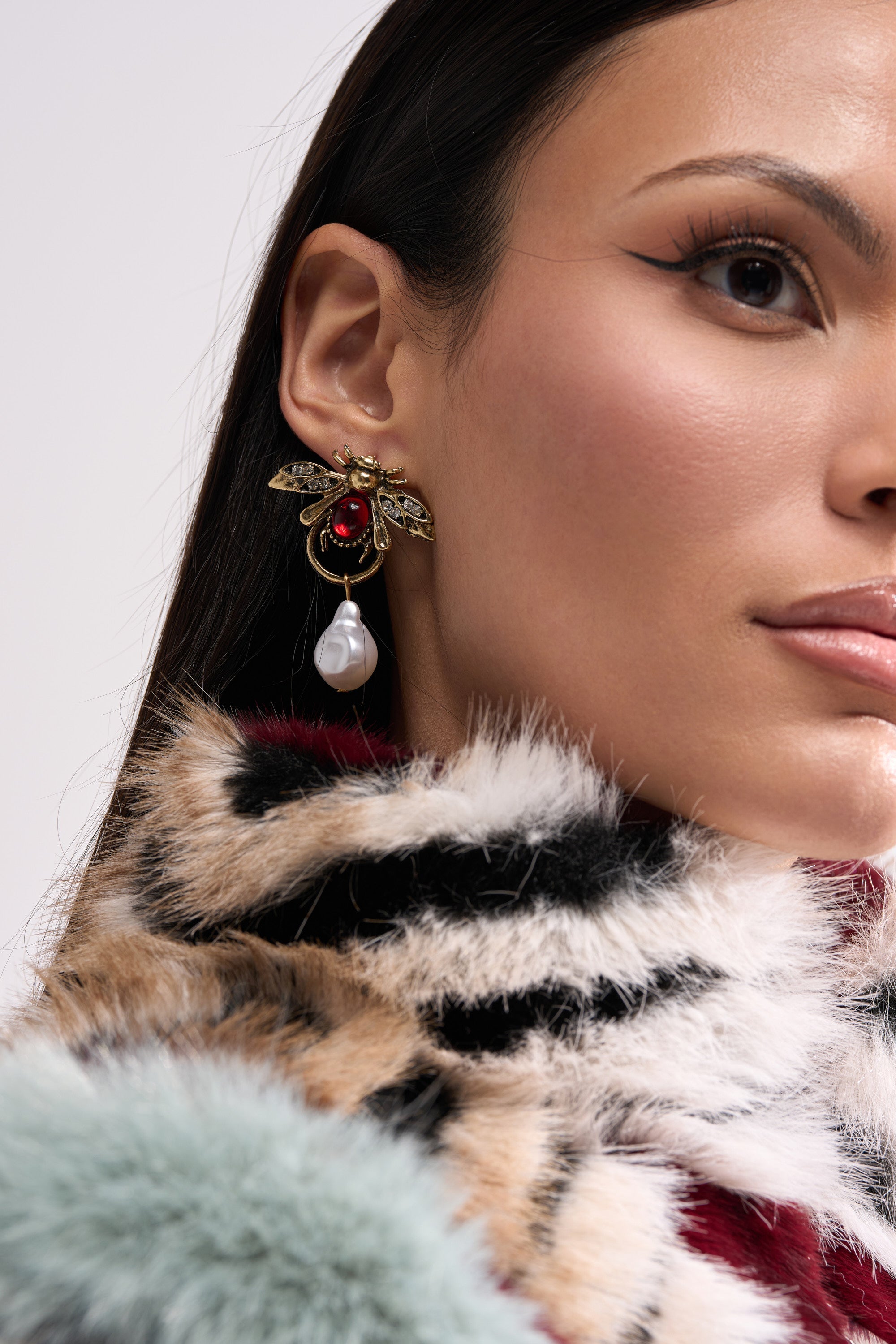 A woman wears a colorful faux fur coat and the OLD MONEY EARRING, featuring a gold bee with a red jewel and dangling faux pearl, in a close-up portrait with sleek hair and winged eyeliner. 1