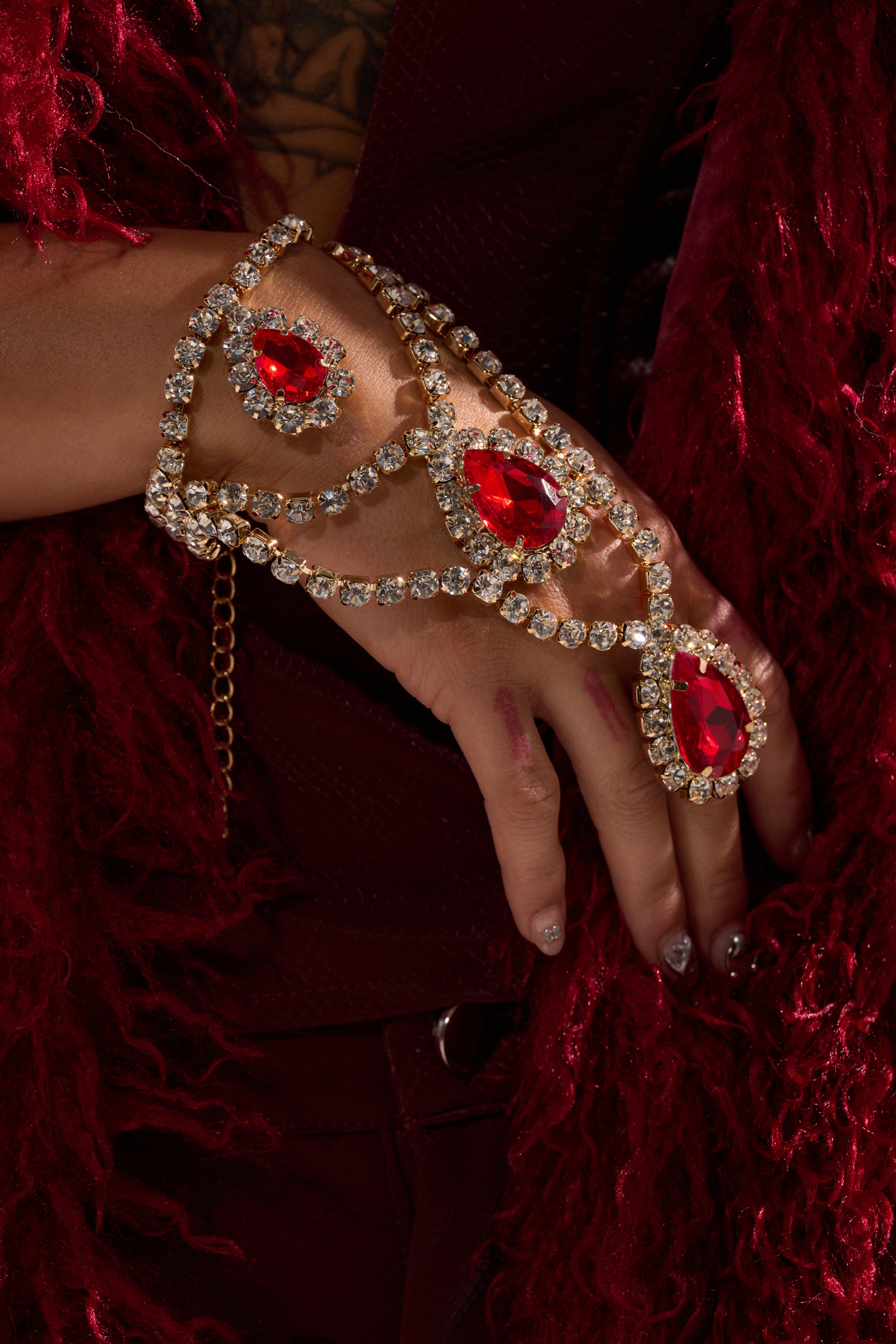 A hand showcases the ELSA BLING RING HAND PANJA, adorned with large ruby red jewels and rows of clear crystals, resting on deep red textured fabric. The wearer’s burgundy attire and a light wrist tattoo are also visible.