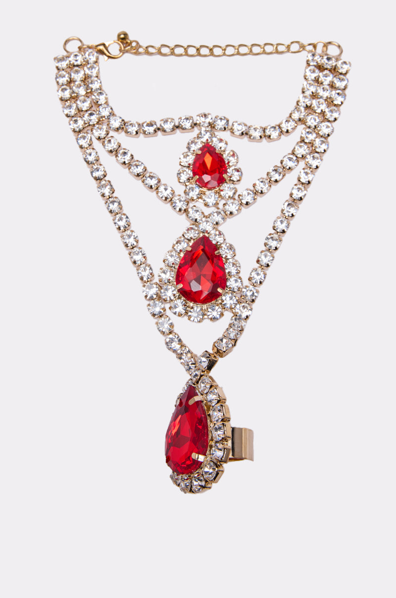 The ELSA BLING RING HAND PANJA is a gold jewelry set with three large teardrop-shaped ruby red gemstones surrounded by clear crystals, linked by gold chains and more crystals. Displayed on a white background, it pairs perfectly with your favorite bling ring.