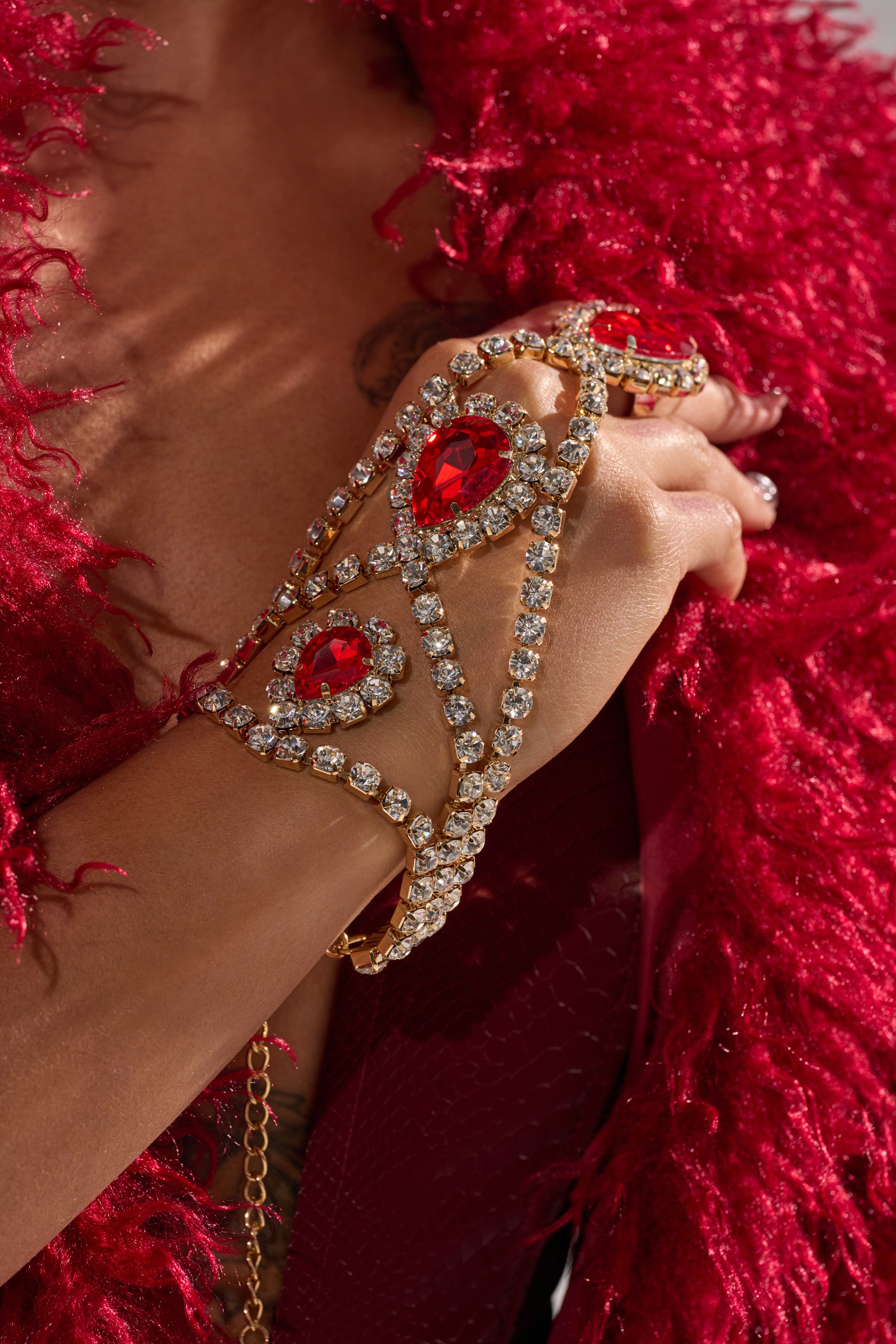 A close-up of a person wearing a red feathered garment, showcasing the ELSA BLING RING HAND PANJA—an ornate ruby red statement bracelet adorned with large gemstones, clear crystals, and matching rings.