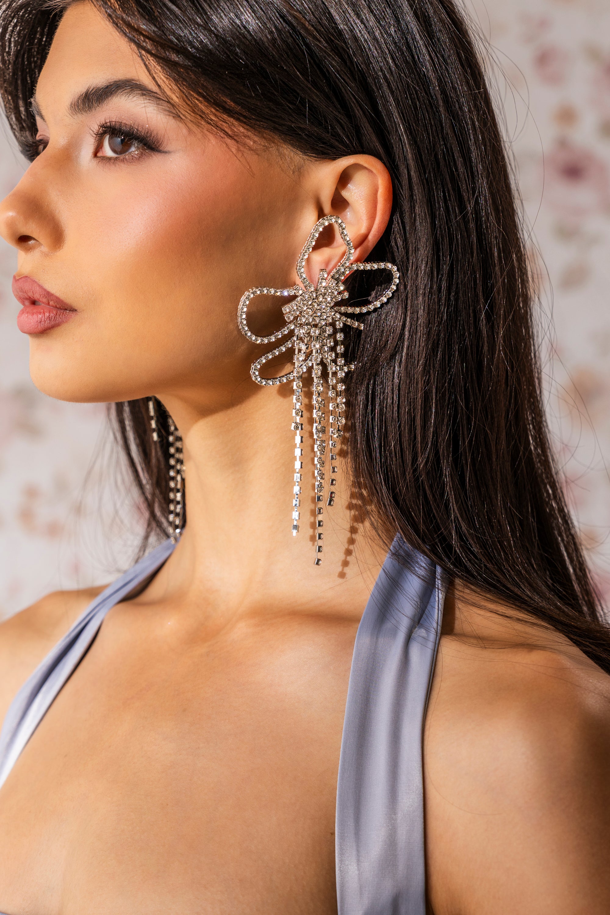 A woman with long dark hair wears the CHECK MY DRIP EARRING, a sparkling rhinestone statement piece with a large flower design and dangling strands. She’s in profile, dressed in a shiny silver-gray top against a floral backdrop.