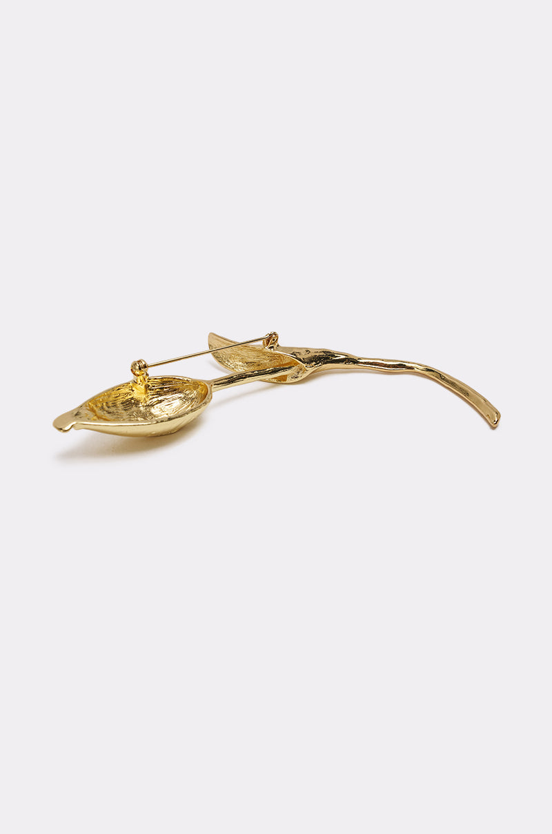 The SPRINGY BROOCH is a sculptural gold-tone pin shaped like a curved branch with a leaf and textured bowl detail, reminiscent of a floral brooch, shown on a plain light background. 3