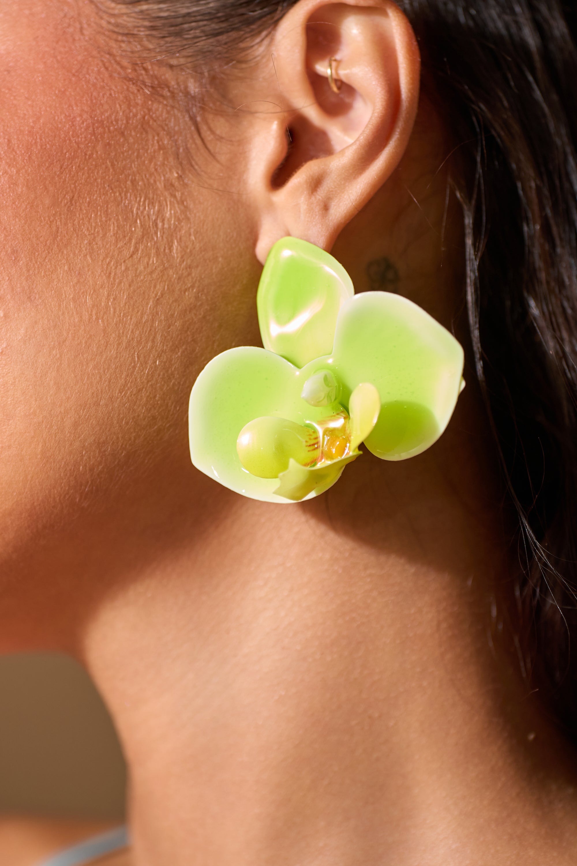 Close-up of a tan-skinned person wearing the TAKE ME OUT EARRING—large, shiny green and yellow orchid-inspired blooms. With dark hair pulled back, these tropical floral earrings stand out beautifully. 1