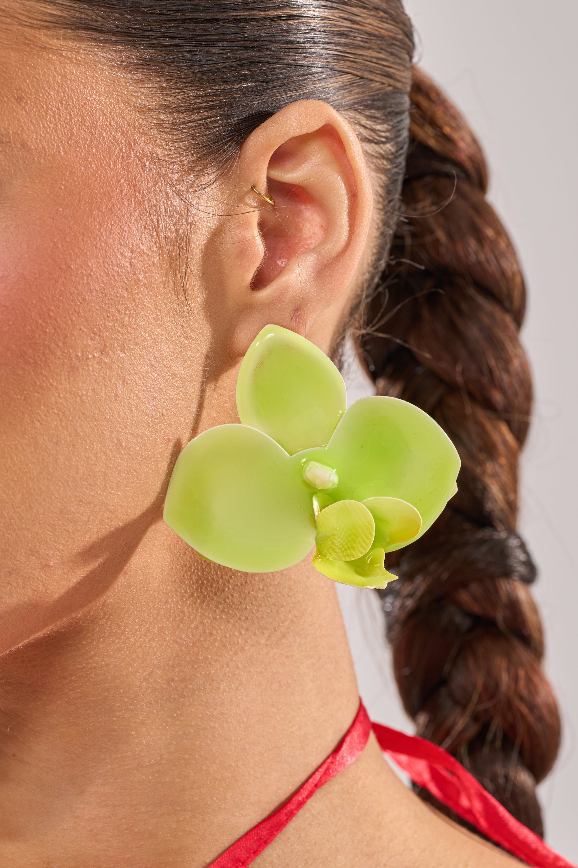 Close-up of a woman's ear wearing the TAKE ME OUT EARRING, a large orchid-inspired lime-green acrylic flower earring. Her dark hair is in a braided ponytail, with a gold ear cuff and red strap around her neck. 6