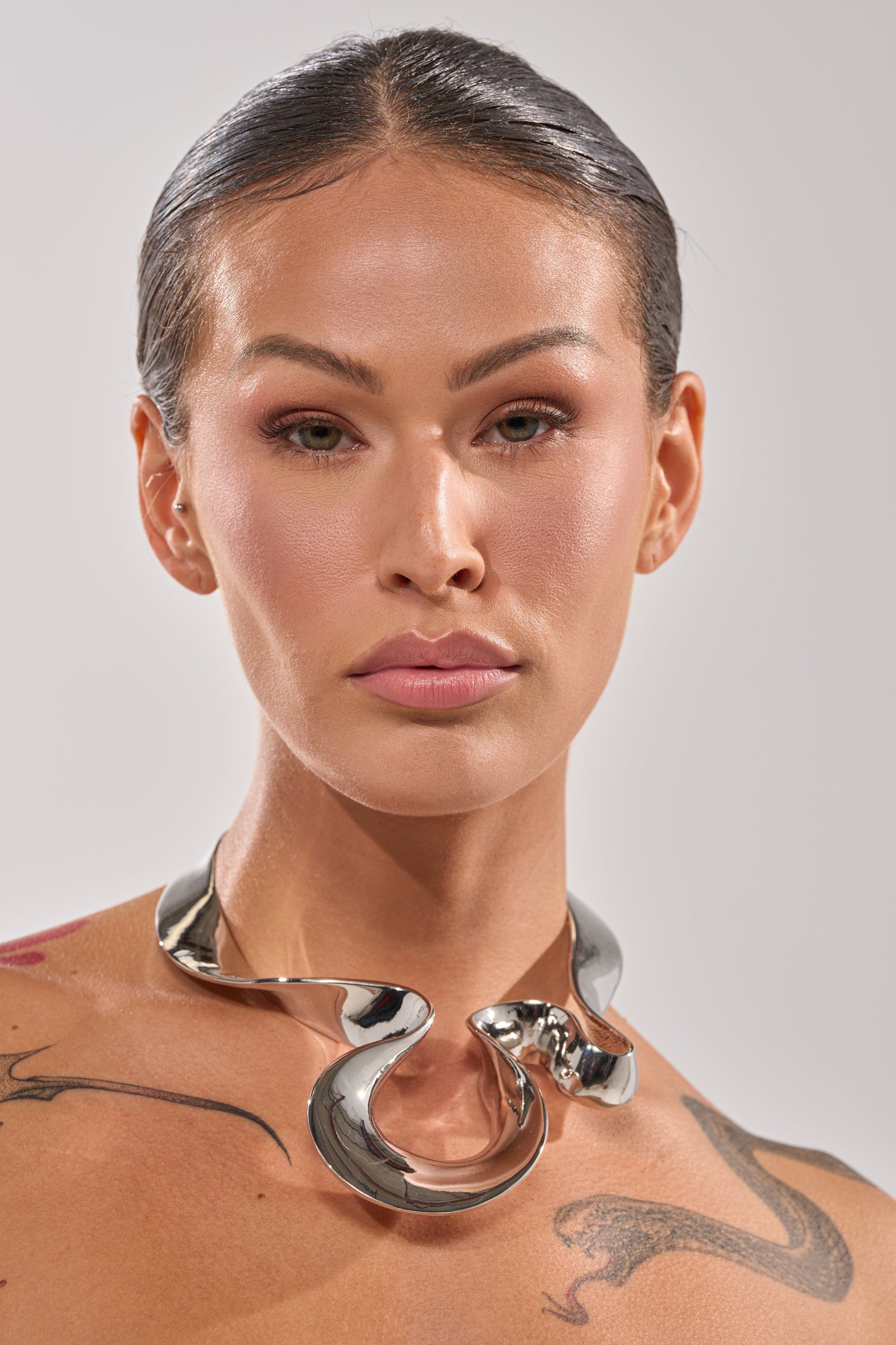A person with slicked-back hair and natural makeup wears the MOLTEN DREAMS NECKLACE, a shiny sculptural silver statement piece. Tattoos are visible on their shoulder and chest against a plain light background. 1
