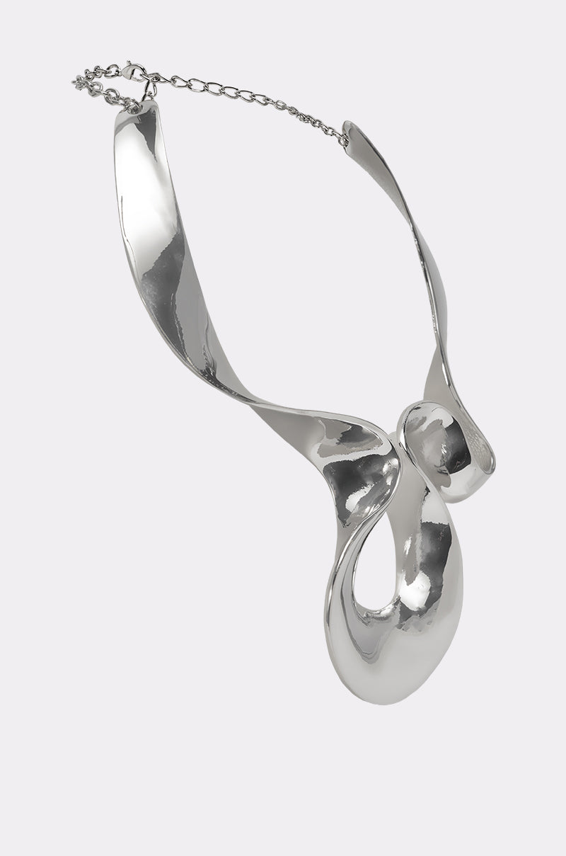 The MOLTEN DREAMS NECKLACE is a modern silver statement piece with a sculptural, abstract design of smooth, flowing curves and reflective surfaces, showcased against a light gray background. 2