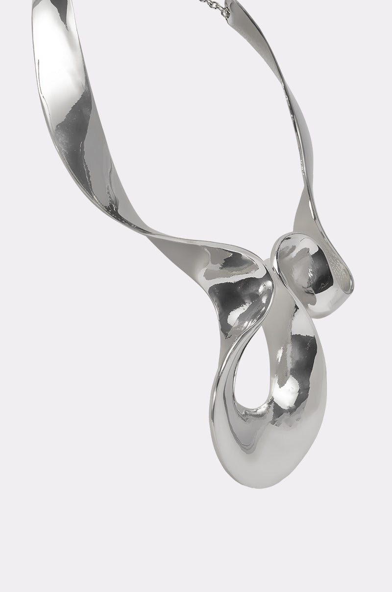 The MOLTEN DREAMS NECKLACE is a sculptural, highly polished silver statement piece with abstract, fluid curves and loops that reflect light on its smooth surface, shown against a plain light gray background. 3