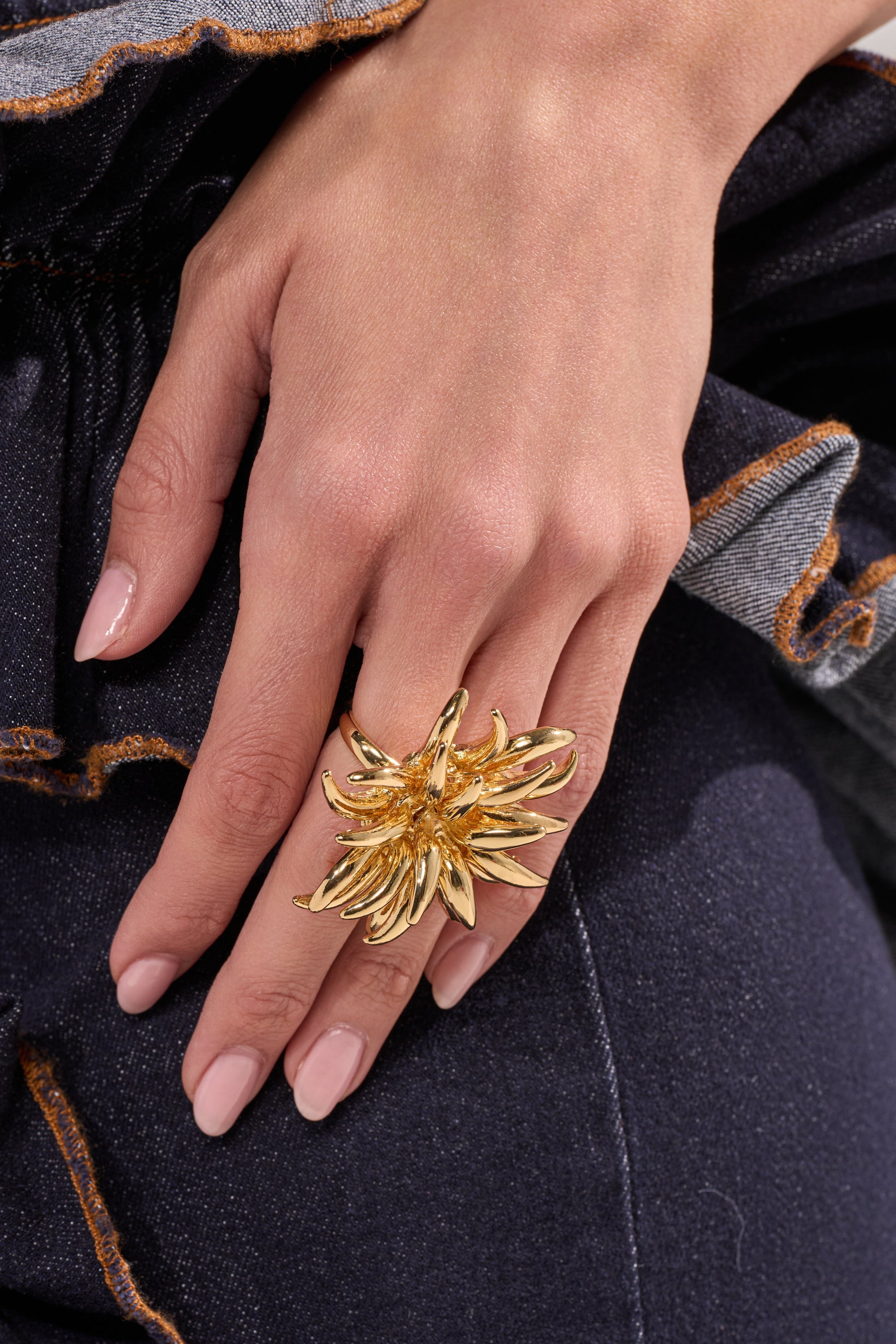 A close-up of a hand with manicured nails on dark denim, wearing the bold SUNBURST GOLD RING—a statement gold piece shaped like a spiked flower. 0
