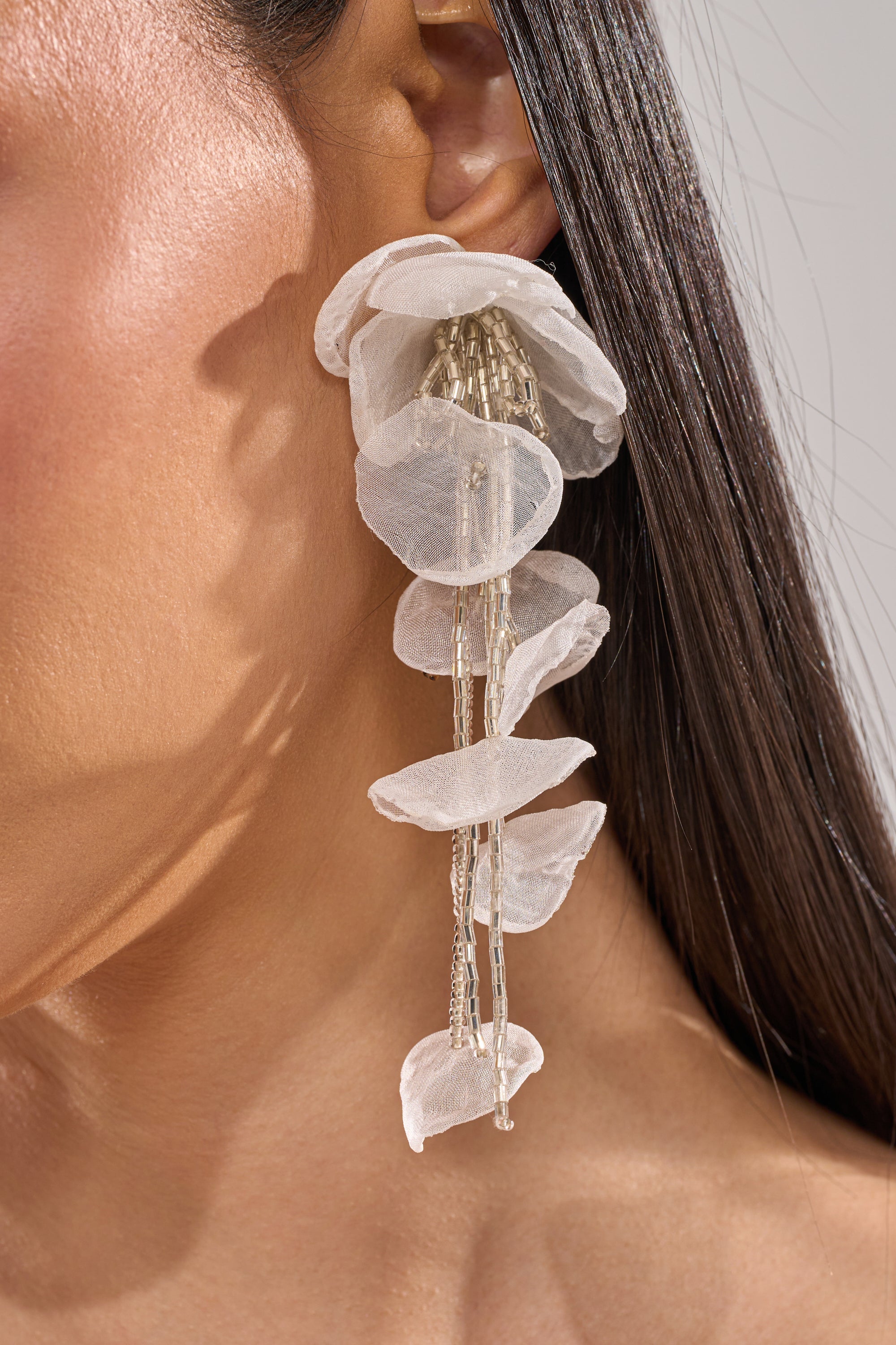 Close-up of a person wearing the IN YOUR DREAMS EARRING—sheer floral drops with translucent, petal-like layers and delicate dangling silver chains—standing out against straight dark hair and smooth skin. 4