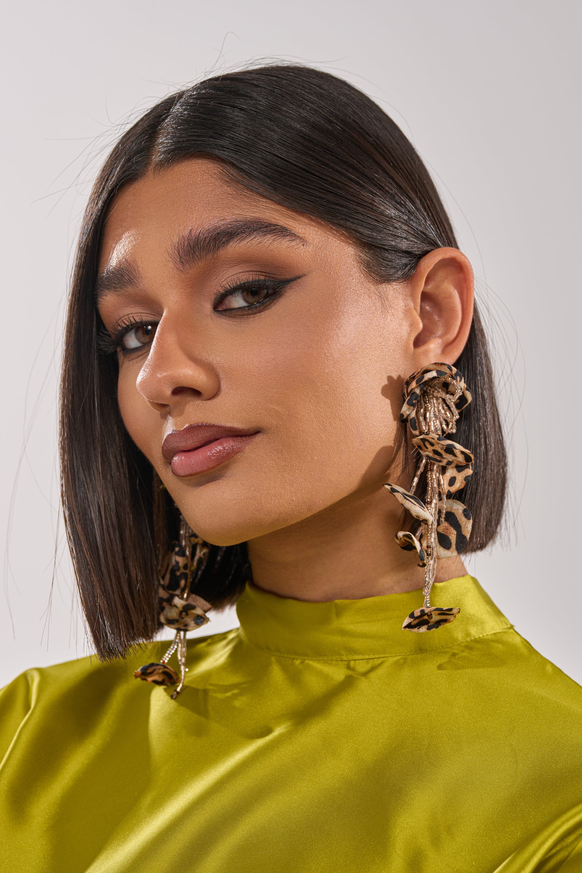 A woman with straight, shoulder-length dark hair wears the bold LEAH EARRING and a shiny lime green high-neck top. She has winged eyeliner, nude lipstick, and gazes confidently at the camera. 1