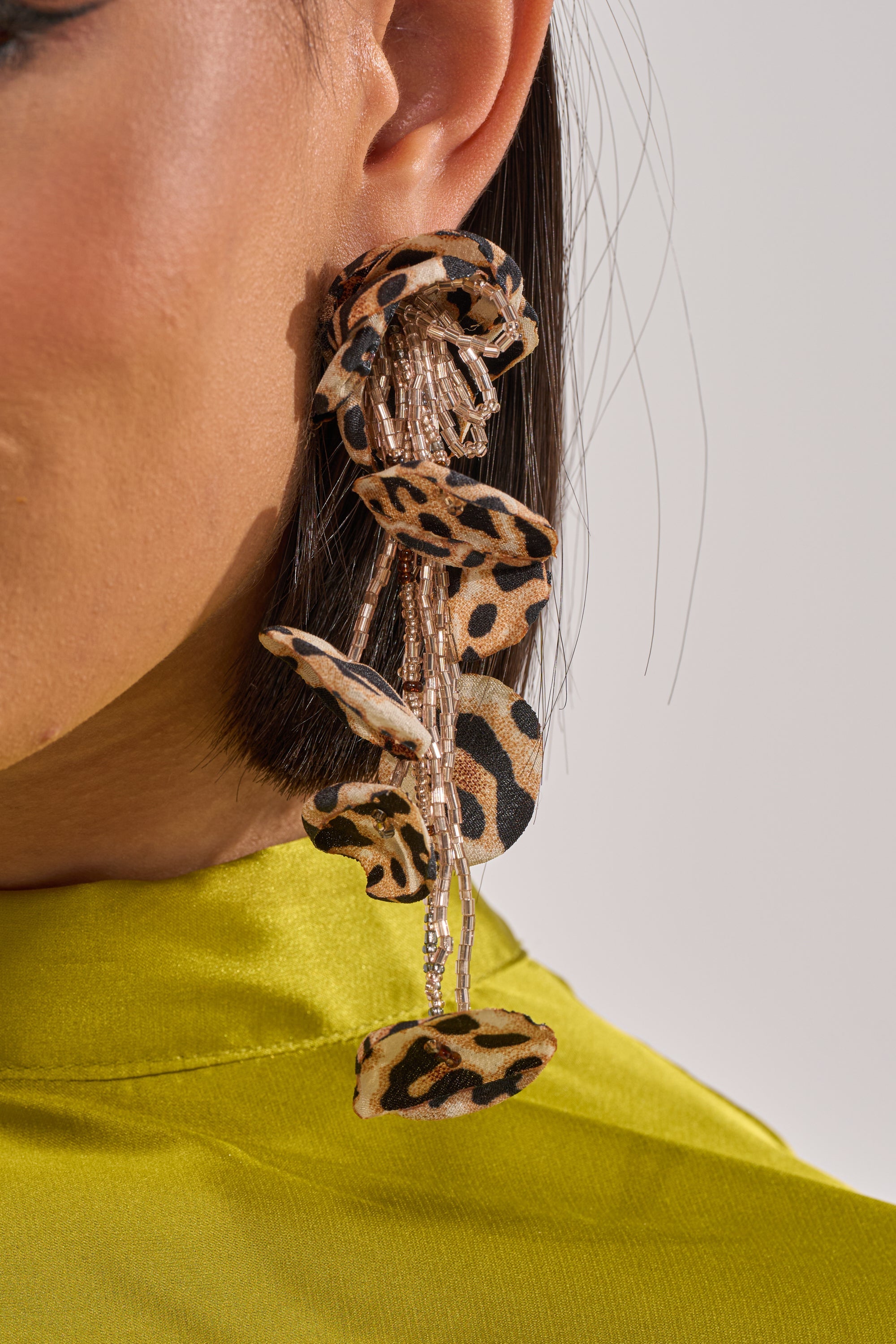 Close-up of a person wearing LEAH EARRING—bold leopard print with beaded fringe—paired with a lime green satin top. Their straight, dark hair frames a visible ear and jawline. 4