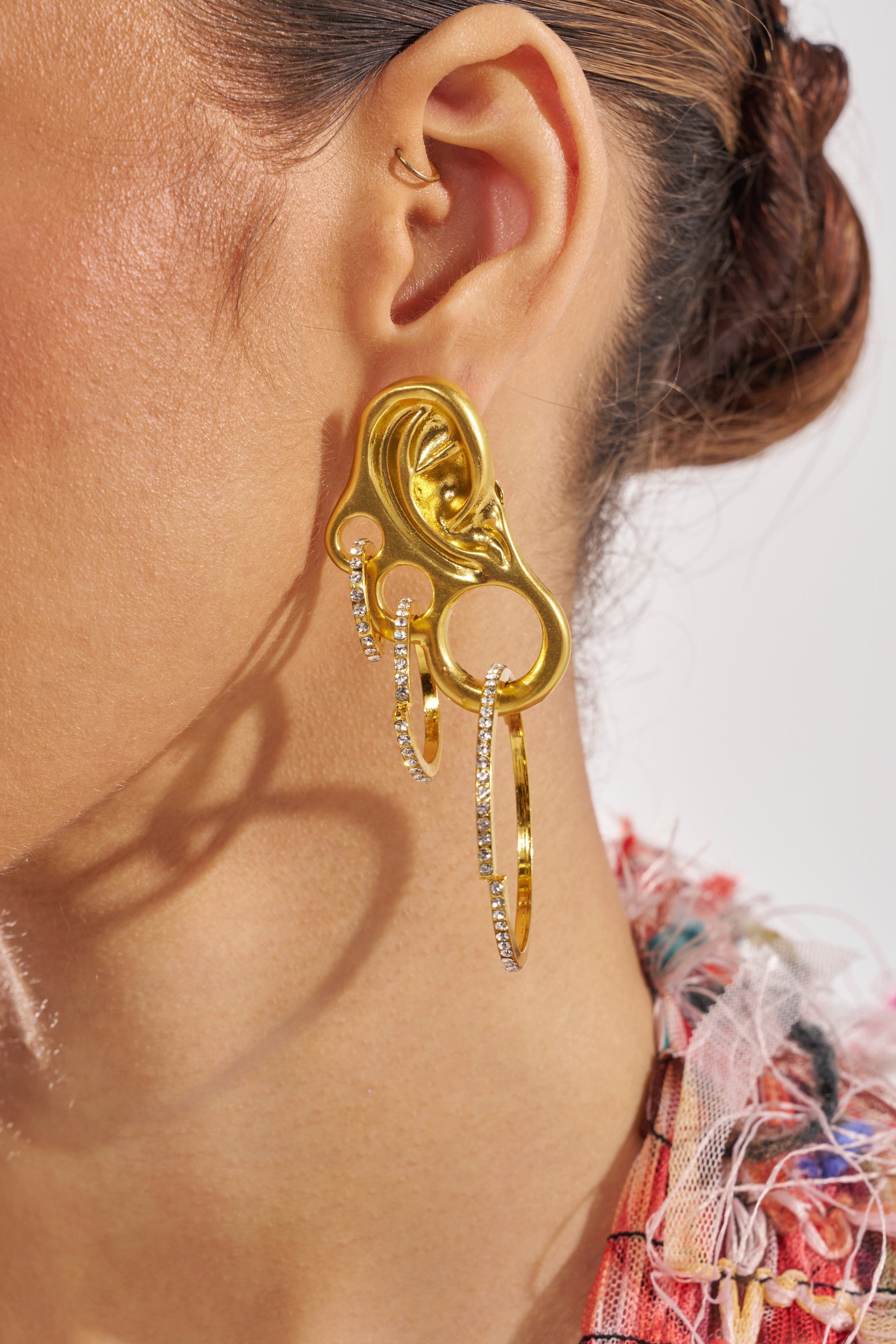 Close-up of a woman's ear wearing the IN MY EAR EARRING, a large, sculptural gold piece with abstract curves and dangling accents, styled with an extra small hoop in her upper ear for a chic layered look. 1